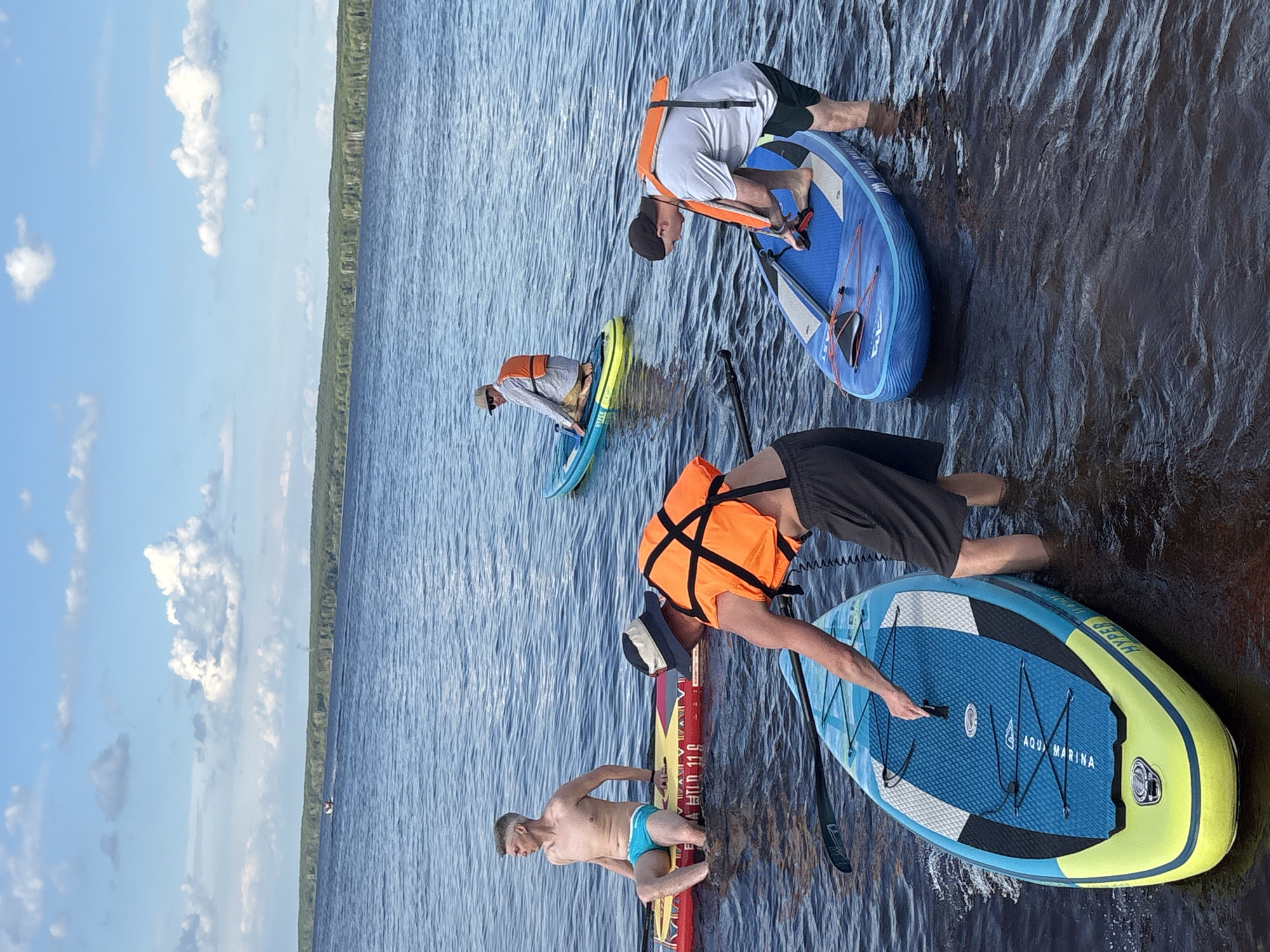 Four individuals are engaged in paddleboarding on a body of water, likely the Kama River in Chaikovsky, Russia, given the context. The scene depicts a sunny day with scattered white clouds in a blue sky. The water is calm and blue, with a wooded shoreline visible in the distance.
In the foreground, one person, wearing an orange life vest and a black baseball cap, is standing in the water and adjusting something on a blue and yellow paddleboard. To their left and slightly behind, another person is seated on a red and yellow paddleboard, wearing blue swim trunks and looking to the left. Further back, a third person, also in an orange life vest and a light-colored hat, is kneeling on a teal and yellow paddleboard. The fourth individual, wearing an orange life vest and a light-colored shirt, is kneeling on a blue paddleboard. All individuals appear to be adults. The paddleboards are various colors and designs, equipped with paddles. The environment suggests an outdoor recreational activity.