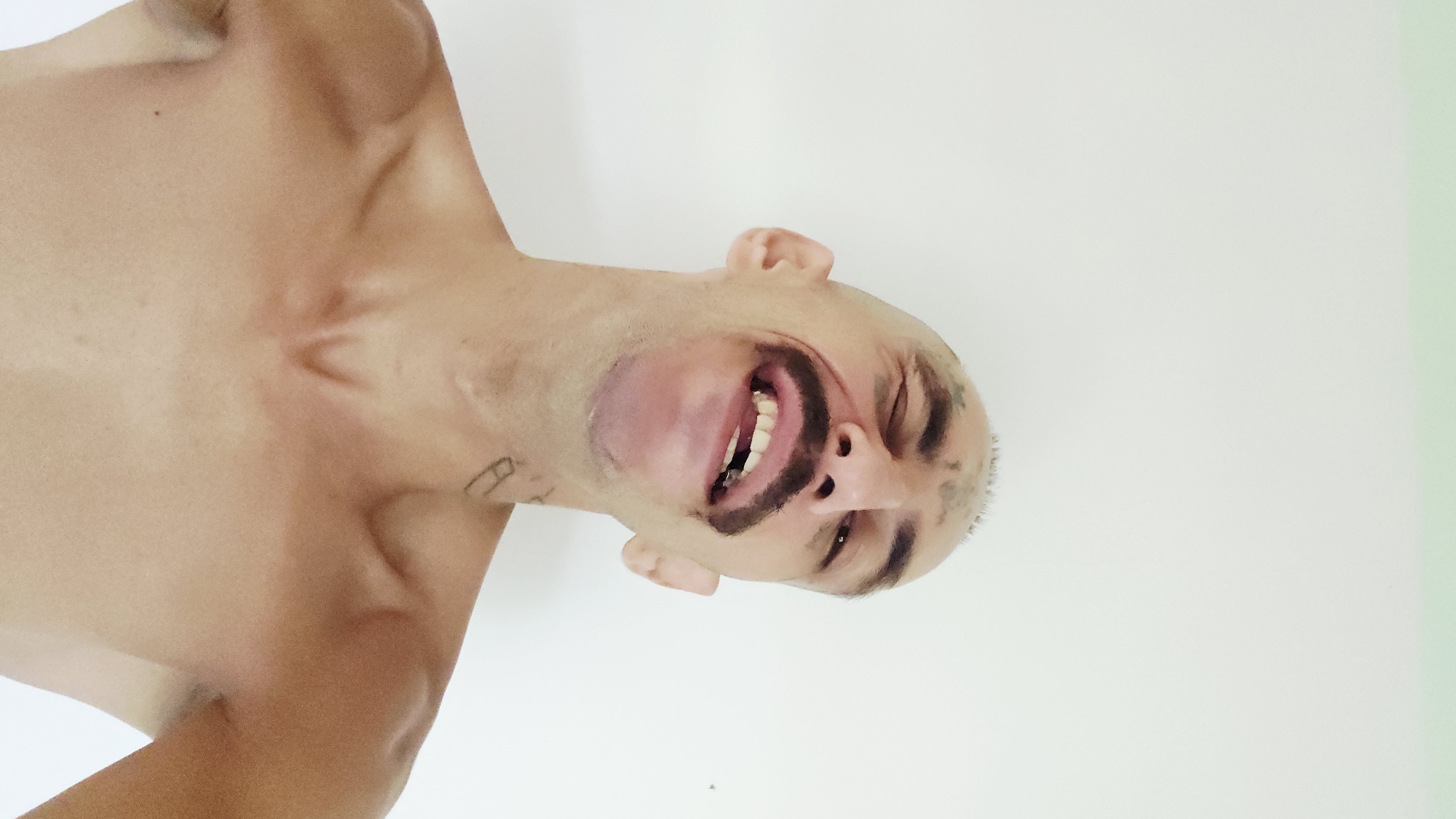 An image, rotated 90 degrees counter-clockwise, depicts a shirtless person from the chest up, against a plain, light-colored background. The individual is fair-skinned, appearing bald or with very closely-shaved hair, and is looking directly at the camera with a wide, open-mouthed smile, revealing both upper and lower teeth.
The most striking feature is the dark face paint applied to the person's face. There is a thick, dark mustache painted above the upper lip and a distinct dark patch resembling a goatee below the lower lip. Fainter dark markings are also visible around the left eyebrow and forehead area. The expression conveyed is one of joy, amusement, or playfulness.
The person's upper torso and shoulders are visible, appearing lean with prominent collarbones. A small, dark, linear tattoo is faintly visible on the left shoulder/upper arm area, resembling a stylized letter "A" or similar symbol. Another faint dark marking, possibly a tattoo, is present on the left side of the neck. The background is uniformly light, suggesting an indoor setting with bright, even lighting.