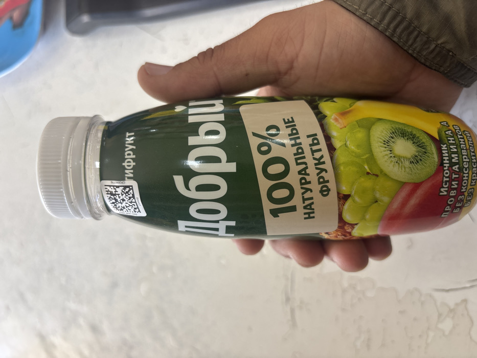 A human hand, appearing to be an adult male's, grasps a green-labeled plastic bottle of "Добрый" (Dobryi) brand fruit juice. The bottle features a white ribbed screw cap and a QR code label with the text "ЦКФРУКТ" near the top. The main label prominently displays "Добрый" in white Cyrillic, followed by "100% НАТУРАЛЬНЫЕ ФРУКТЫ" (100% NATURAL FRUITS). Visuals on the label depict various fruits, including green grapes, a sliced kiwi, a banana, and a red fruit, suggesting a multifruit blend. Additional text states "ИСТОЧНИК ПРОВИТАМИНА А БЕЗ КОНСЕРВАНТОВ БЕЗ КРАСИТЕЛЕЙ" (SOURCE OF PROVITAMIN A WITHOUT PRESERVATIVES WITHOUT DYES). The hand, with a visible light-colored sleeve, holds the bottle from the right side. The background is an out-of-focus, light-toned surface, possibly a table or countertop, with a blurred blue object and a dark, indistinct item in the upper right quadrant. No specific visual cues within the image directly indicate the precise location within the city of Lehvaz, Armenia.