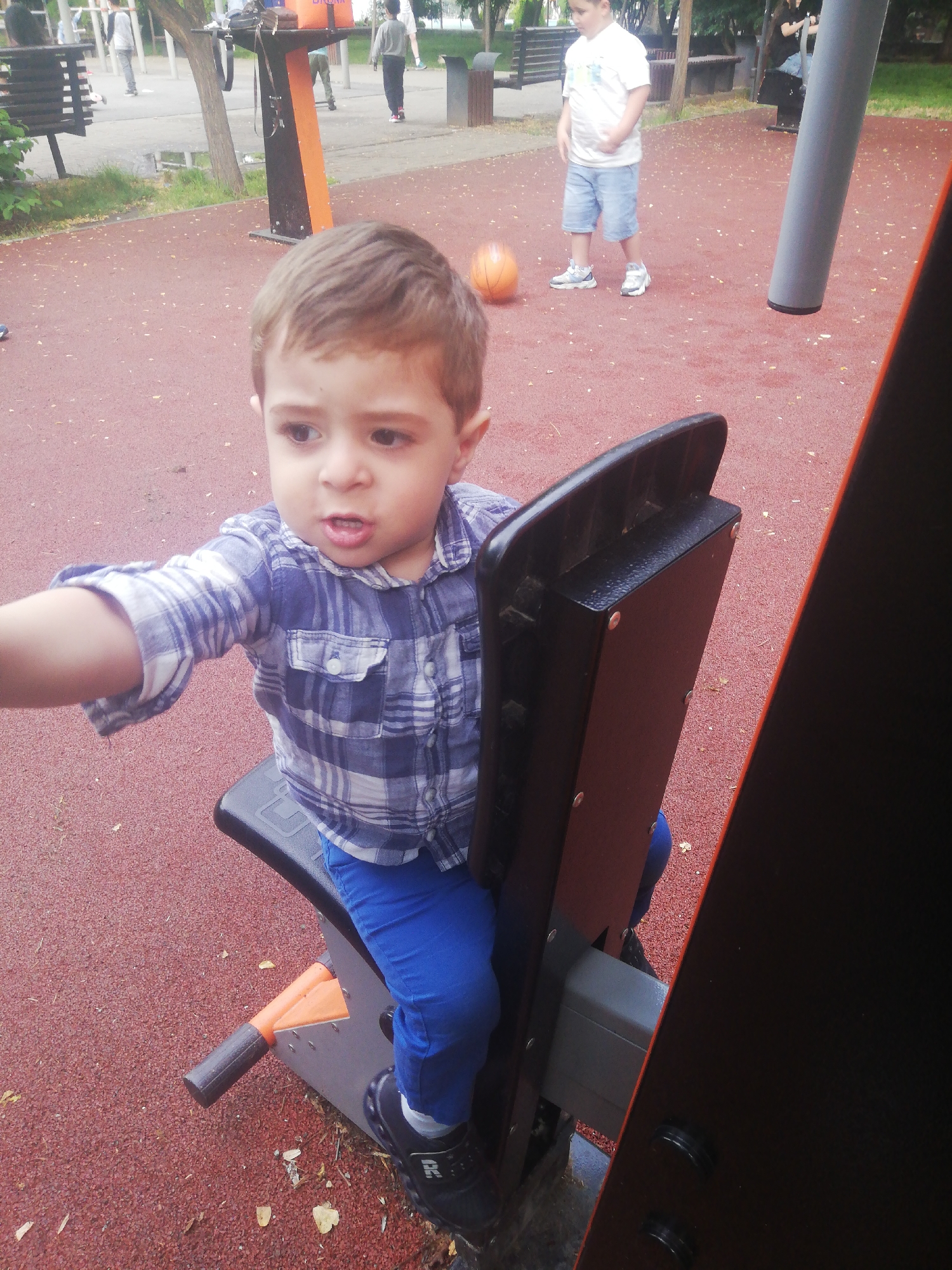 The image shows a young boy sitting on a black and orange playground seat. He is wearing a blue and white plaid shirt and blue pants. His left arm is extended towards the camera. The playground has a red rubberized surface. In the background, there are other children and adults, as well as trees and playground equipment. This scene appears to be a public park or playground. The location is in Yerevan, Armenia, as indicated by context not present in the image itself.