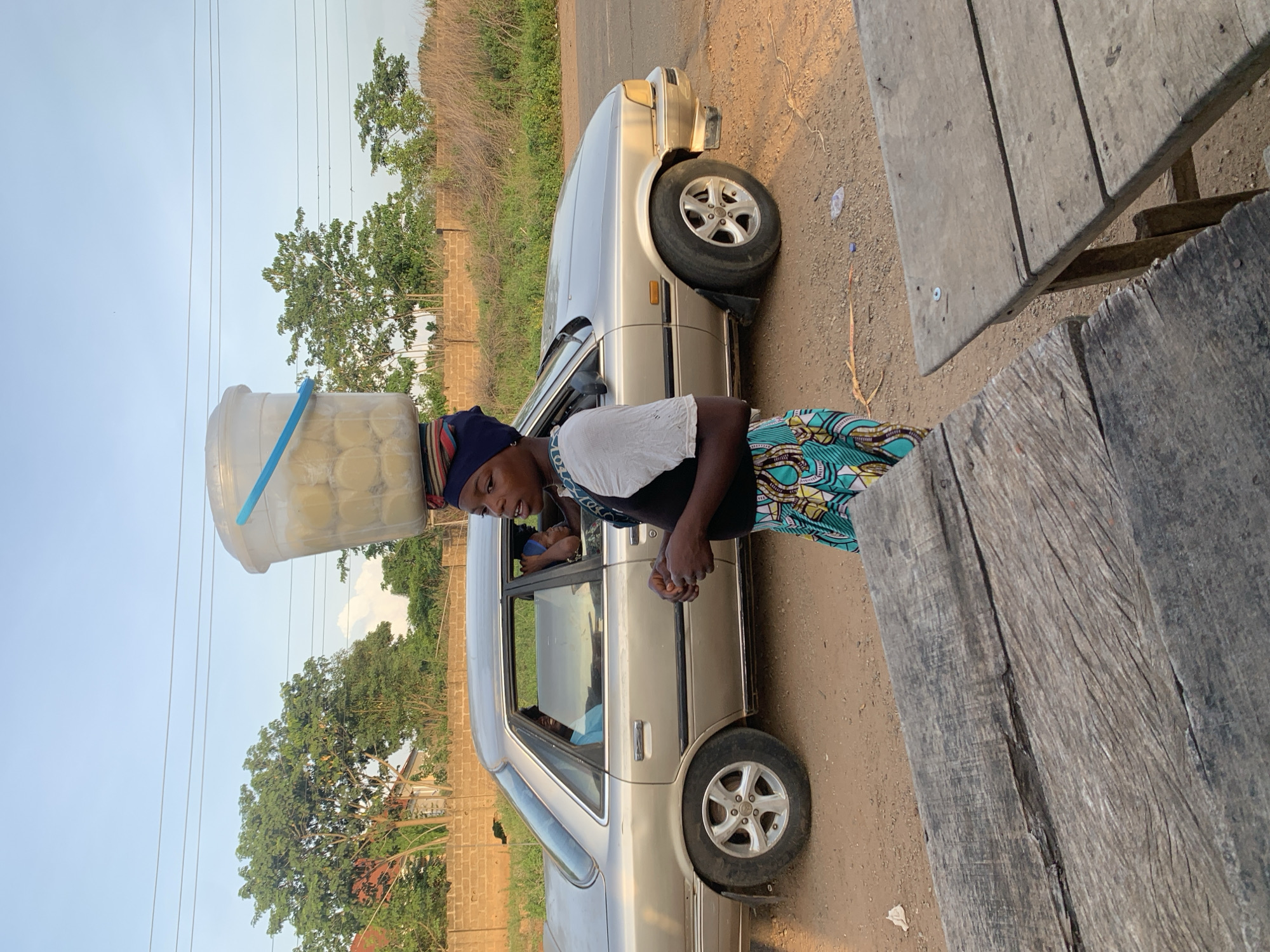 A woman carries a container of what appears to be cheese curds on her head next to a parked silver sedan, possibly a Toyota, in Gbeleko, Nigeria. She is wearing a patterned skirt, a white shirt, and a black vest. A person is visible in the back seat of the car. A rough wooden table is visible in the lower right corner of the image. The area has trees and a partly cloudy sky. Electrical wires run overhead. The ground is dusty. A brick wall is in the distance.