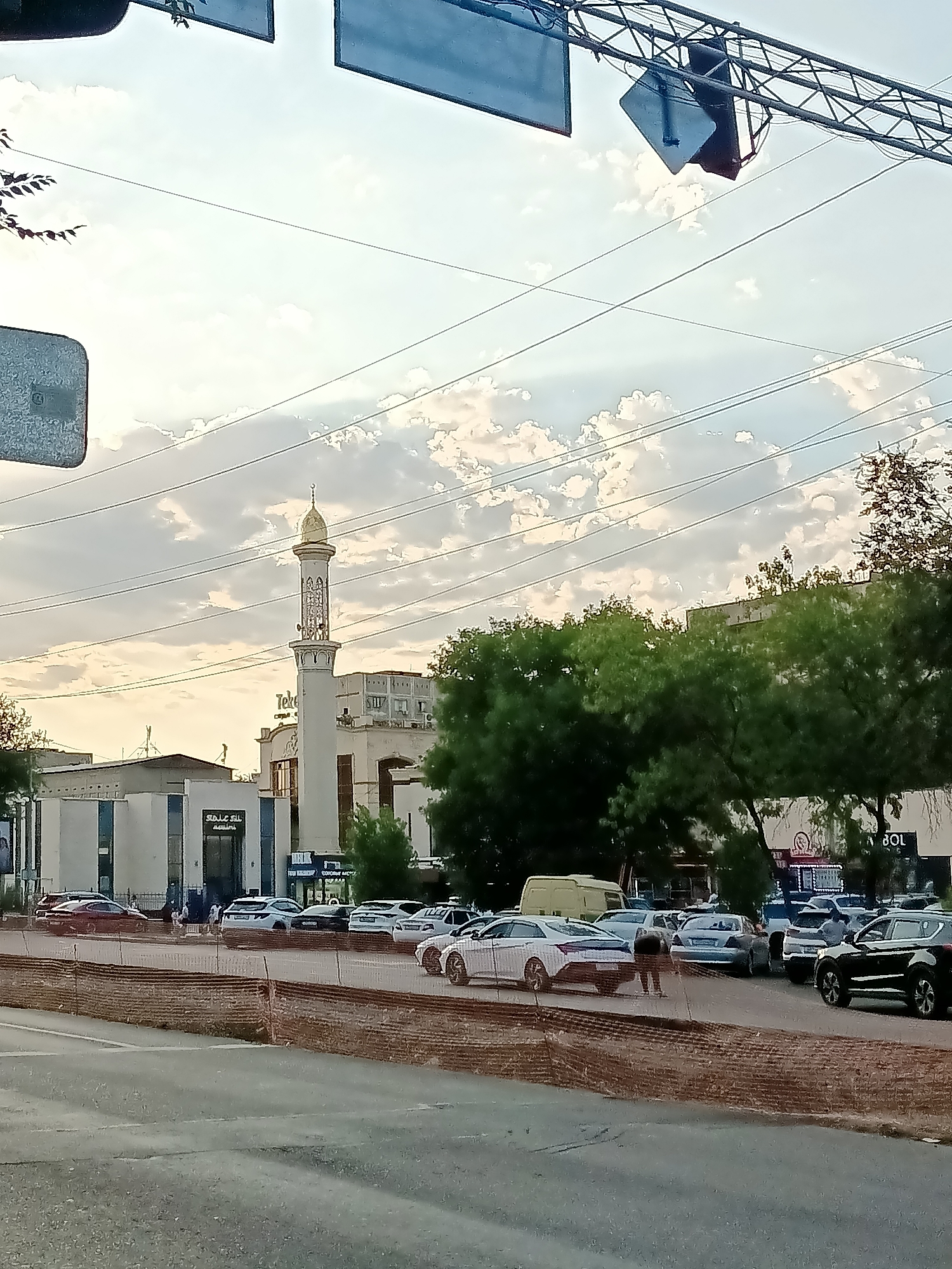 In Almaty, Kazakhstan, a mosque with a tall, slender minaret topped with a golden dome stands prominently against a cloudy sky. The mosque features white walls with blue accents. In front of the mosque, a paved area serves as a parking lot filled with various vehicles, including cars and a yellow van. Some people are visible in the parking area, some standing, others walking. The scene is bordered by a low, rust-colored brick wall with a mesh fence. Trees line the background, obscuring some of the buildings behind them. Overhead, power lines crisscross the sky, and traffic lights are suspended above the roadway.