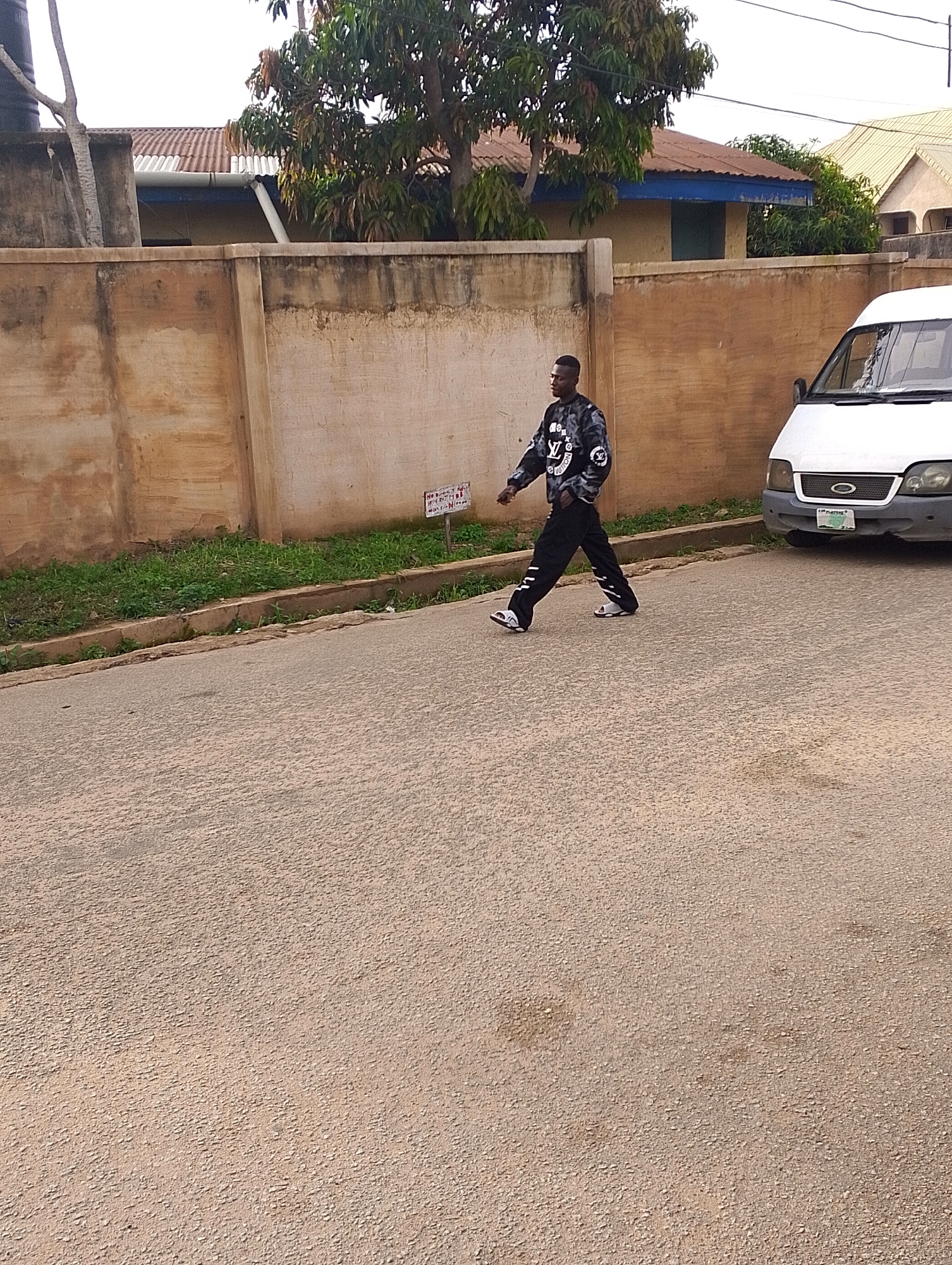 A young Black male wearing a black sweatshirt with a white LV logo pattern and black pants with white stripes walks on a paved road in Jos, Nigeria. He is on the left side of the frame, facing right and slightly away from the camera. A white van is parked on the right side of the road, partially visible. Behind the road is a concrete wall, and behind the wall are buildings with corrugated metal roofs and trees. The sky is overcast.