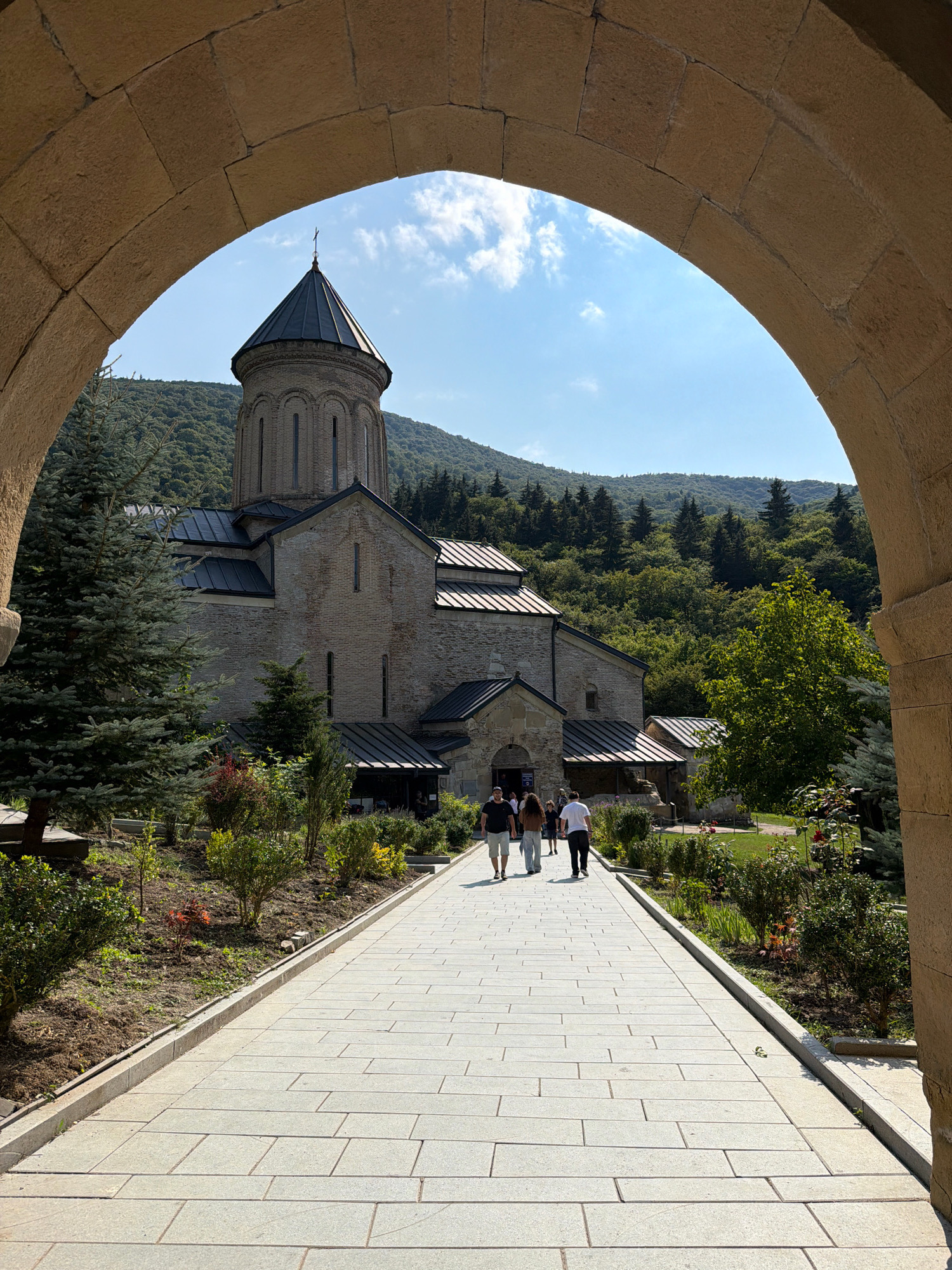 The image shows an arched stone entranceway framing a view of the Gremi architectural complex in Krobani, Georgia. A paved walkway leads towards a stone church with a dark, conical roof and a cross atop it. Green trees and lush vegetation surround the church, with a hilly, tree-covered landscape forming the backdrop under a bright blue sky with scattered clouds. Several individuals, appearing to be tourists, walk along the pathway towards the church. The scene is bathed in natural daylight, suggesting it was captured during the daytime.