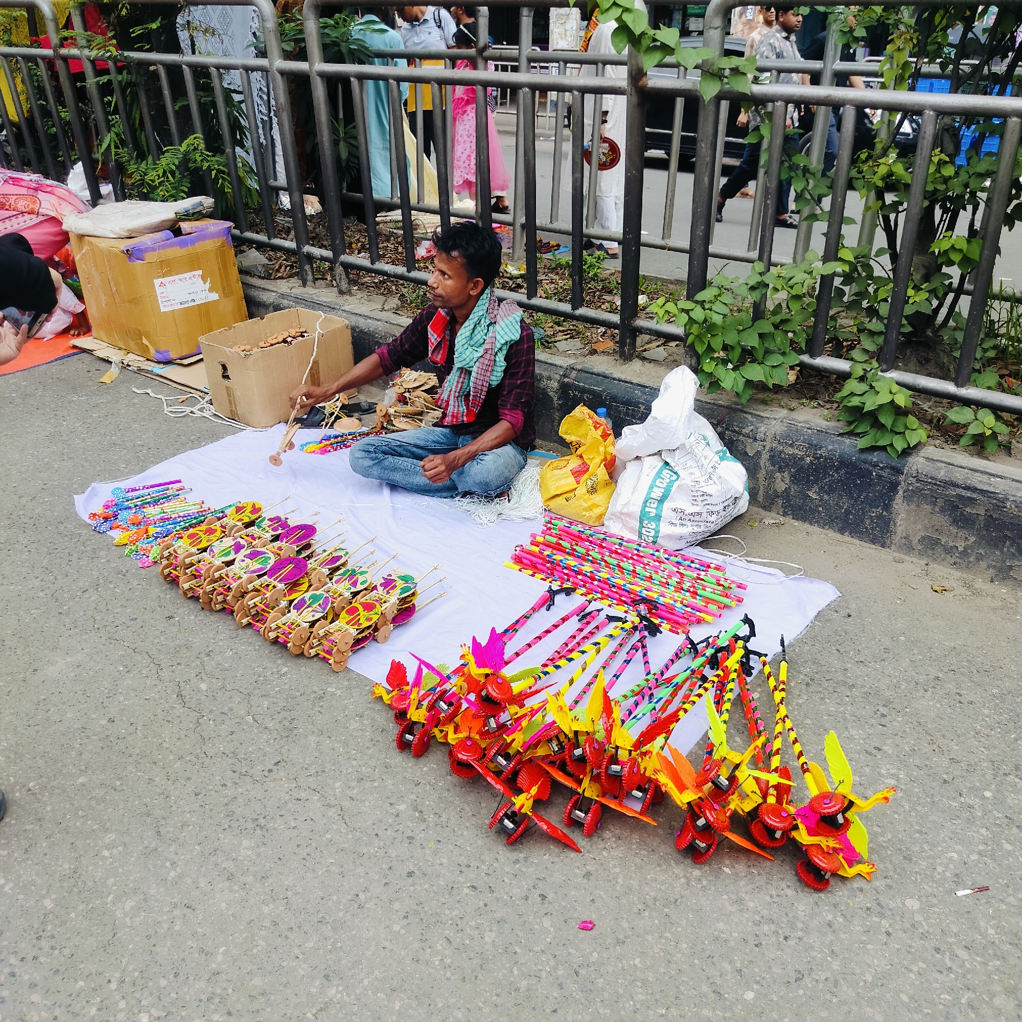 An outdoor daytime shot shows a street vendor in Dhaka, Bangladesh. The vendor is a young man sitting cross-legged on a white cloth, surrounded by colorful handmade toys displayed for sale. He is wearing a maroon plaid shirt, blue jeans, and a light blue and red scarf. The toys include wooden cars, windmills, and bird-shaped decorations attached to sticks. The vendor is holding one of the wooden cars, appearing to be assembling or adjusting it. Behind him are a cardboard box, a yellow plastic bag, and a white cloth bag. The background features a metal railing with greenery, people walking, and vehicles moving on the street. The scene appears to be a busy public area, likely a sidewalk adjacent to a road.