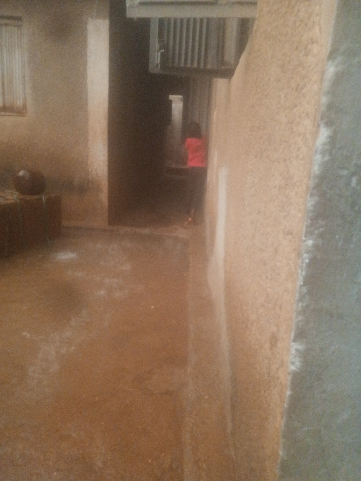 This image captures a narrow outdoor passage between buildings during rainfall. The ground is wet and appears muddy or covered in standing water, with visible splashes indicating ongoing precipitation.
On the left side of the frame, a textured light brown wall features a window covered by vertical blinds or slats. Below the window, several dark, rectangular objects are stacked, possibly bricks or blocks, with a round, dark object resting on top. The wall opposite, on the right, is also textured and a similar light brown color.
Further into the passage, in the mid-ground, a person is visible from behind, wearing a red top and dark pants, walking away from the viewer. They are partially obscured by the shadows and distance. Above the person and in the upper background, there is a metallic structure with vertical elements, possibly part of an overhead walkway, a building's exterior, or industrial equipment.
The overall lighting suggests daytime, likely overcast due to the rain. The environment appears functional and somewhat rustic, characteristic of an urban or semi-urban setting. No specific text is visible in the image. The image quality is somewhat grainy, which affects the clarity of distant details.