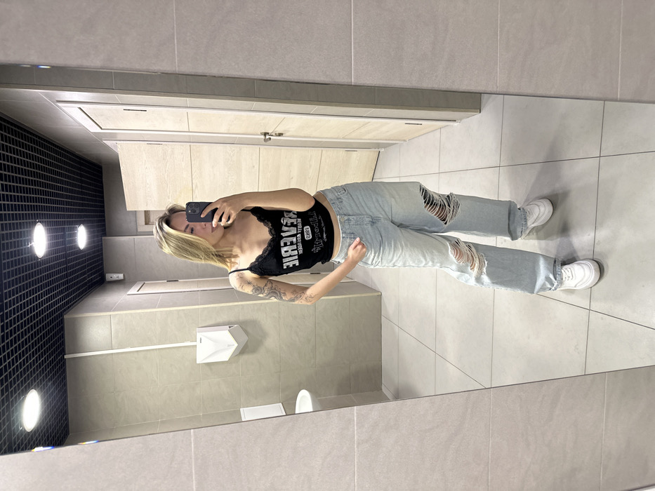 The image displays a female individual taking a mirror selfie inside a public restroom. The female, with blonde, shoulder-length hair, is depicted from the mid-thigh up, holding a black smartphone with her right hand to capture her reflection. She wears a black spaghetti-strap crop top with lace trim and the reversed text "REVERIE BEAUTY ACADEMY 1985." Her attire includes light-wash, wide-leg ripped jeans featuring large tears on both knees, and white athletic shoes. A dark, possibly dragon-themed, tattoo is visible on her left arm.
The restroom features light-grey rectangular tiled walls and lighter grey square tiled floors. The ceiling is a black grid system containing multiple circular recessed light fixtures. Visible fixtures include a white wall-mounted hand dryer, a white sink basin, a portion of a white toilet, and two light-colored wooden or laminate stall doors. A thin white conduit runs vertically along the left wall, and a small white sensor-like box is mounted high on the wall to the right of the hand dryer. The specific location within Khimki, Russia, cannot be determined from the visual information provided.