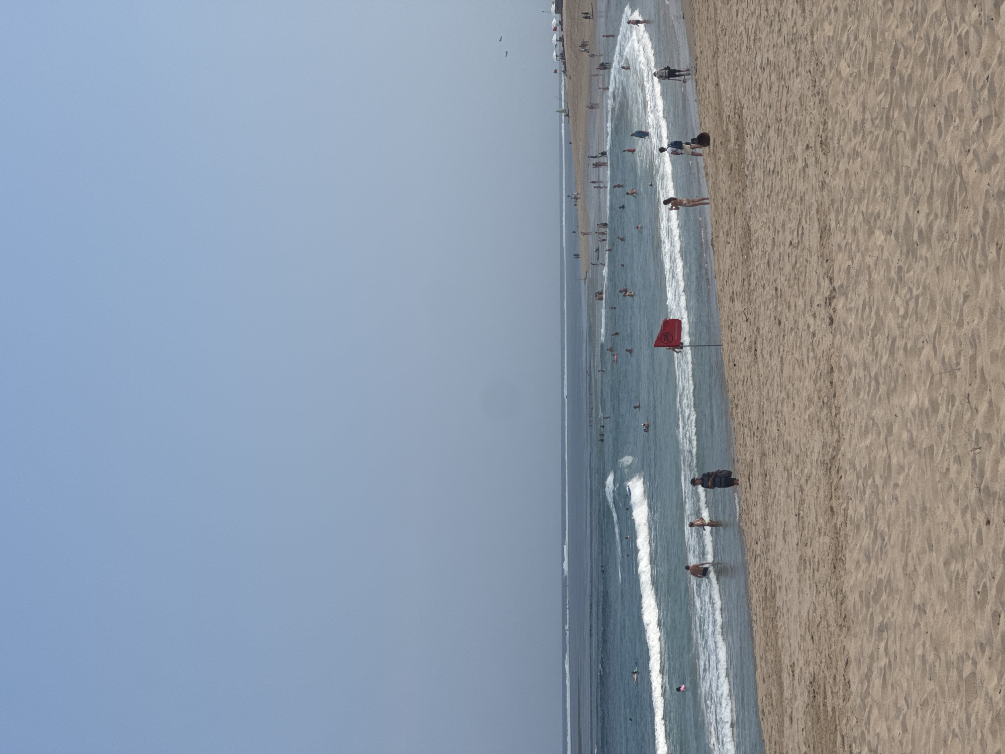 This image depicts a crowded beach scene in Kuta Selatan, Indonesia, under a clear, hazy sky. The foreground is dominated by a wide expanse of light brown sand. In the middle ground, the turquoise ocean has moderate waves rolling towards the shore. Numerous people are visible in the water, some swimming, others surfing on small boards. Along the waterline and on the sand, more people are scattered, walking, sitting, or standing. A bright red flag is planted in the sand near the water's edge, likely indicating a warning or a designated swimming area. The background shows the vast ocean meeting the horizon, with a few distant white structures resembling umbrellas or tents on the far beach. Two small dark shapes, possibly birds, are visible in the upper right quadrant of the sky.