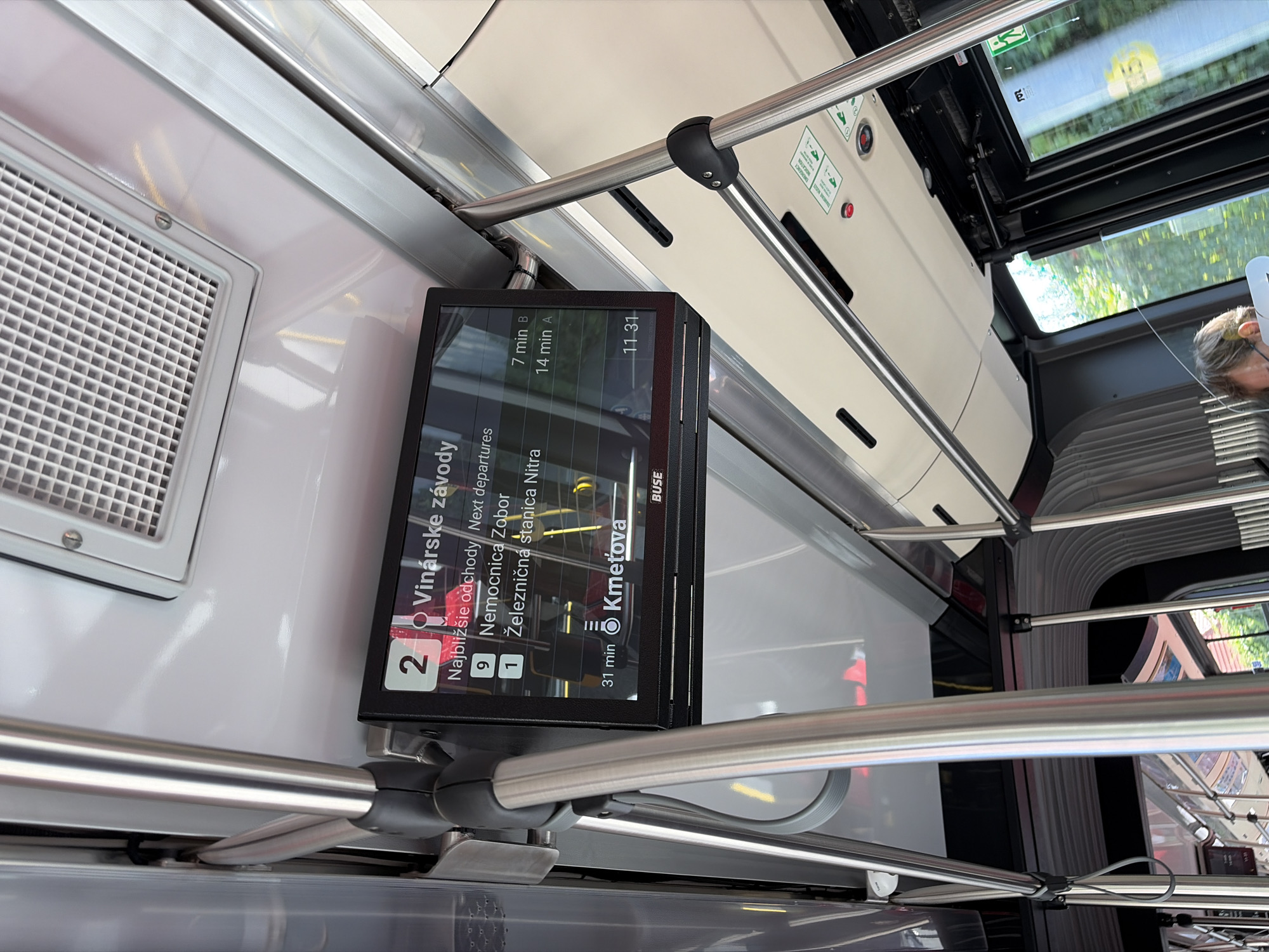 The image shows the interior of a public transport vehicle, likely a bus, in Nitra, Slovakia. A digital display screen is prominently featured, showing "Vinárske závody" which translates to "Wineyards factories." Below this heading, it lists "Najbližšie odchody Next departures" followed by departure information for bus routes 9 and 1. Bus route 9 is heading to "Nemocnica Zobor" and route 1 to "Železničná stanica Nitra" (Nitra Railway Station). Times for departures are listed as 7 minutes and 14 minutes, with designations "B" and "A" respectively. Another time, 11:31, is also visible. At the bottom of the screen, "Kmet'ova" is displayed with a time of "31 min", suggesting a destination or stop with an estimated arrival time. The screen is branded "BUSE".
Several metal poles, typical of public transport interiors, are visible, along with the ceiling paneling and lighting. The environment suggests the vehicle is either stationary or moving, with the exterior seen through the windows in a blurred, motion-effected view, showing greenery and trees. The overall scene is clean and functional, typical of modern public transportation.