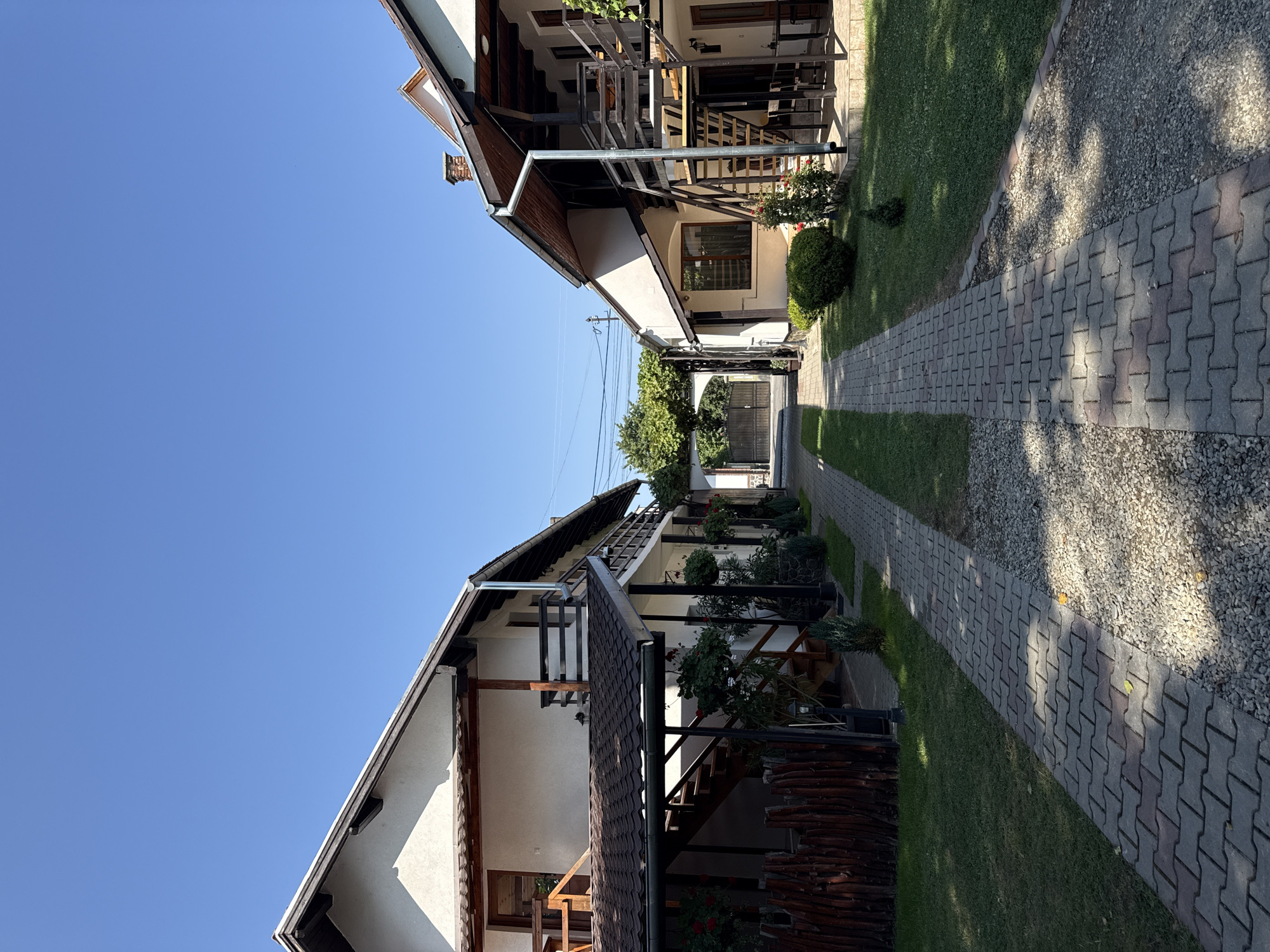 A vertical, eye-level shot shows a pathway leading to a gated archway, flanked by two white, two-story buildings with dark wood accents. The pathway is paved with interlocking grey and reddish-brown bricks, bordered by green grass on the left and gravel on the right. Both buildings feature dark wood balconies, staircases, and roof overhangs. The left building has a wooden fence in front of it. The right building has outdoor seating under its balcony. The sky is a clear, bright blue. The scene is likely a guesthouse or private residence in Cârțișoara, Romania, given the architectural style and the presence of outdoor amenities suggesting a tourist-oriented location.