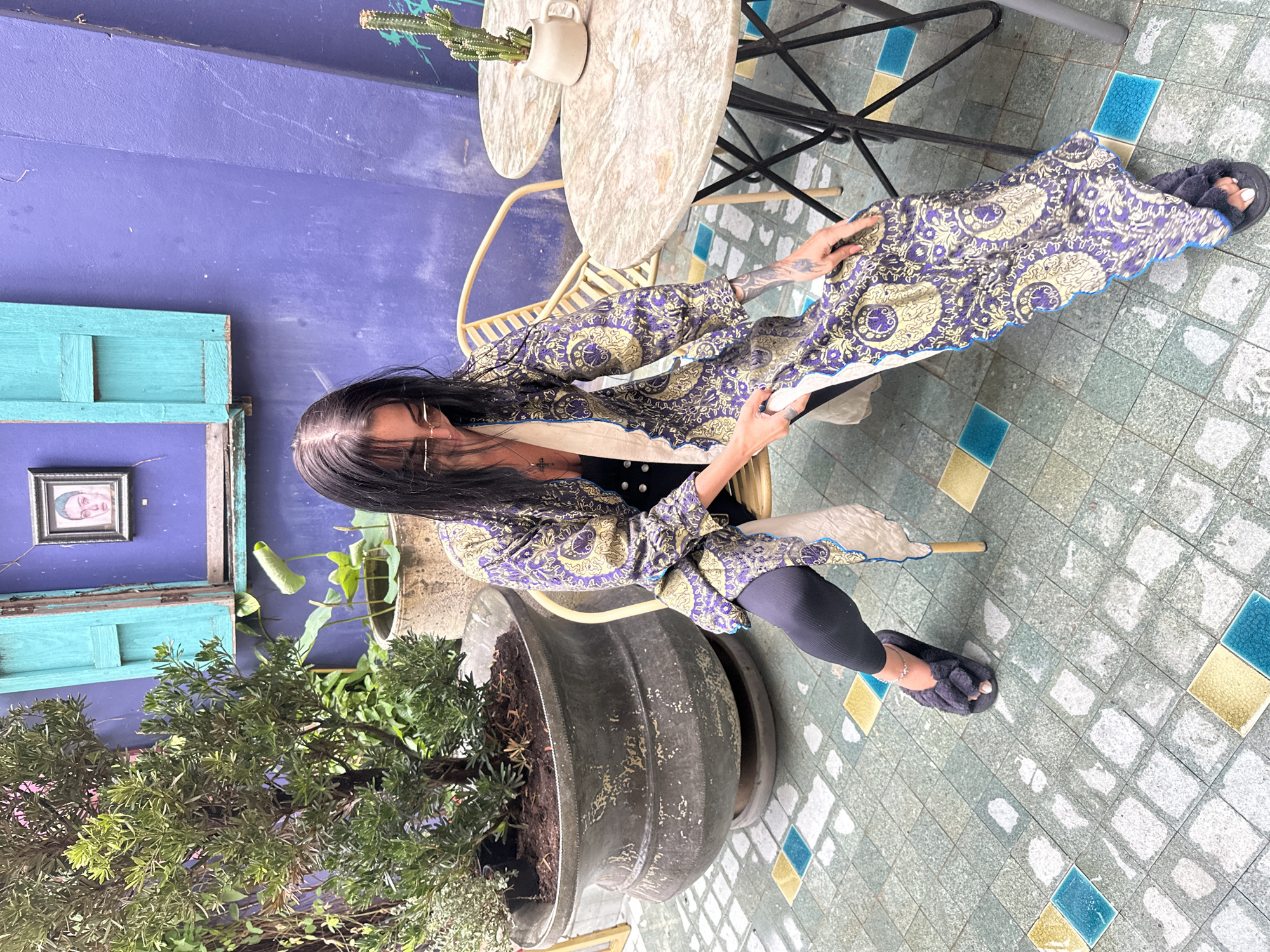 The image depicts an outdoor patio in Baan Lipa Noi, Thailand. A woman with long dark hair, wearing sunglasses, a black top, black leggings, and dark fuzzy slippers, is seated on a light yellow woven chair. She wears a long, open jacket with a purple and gold intricate paisley pattern and a blue trim, which she is adjusting with both hands, looking downwards. A cross pendant hangs from her neck, and a delicate anklet adorns her right ankle. Tattoos are visible on her left forearm.
The background features a vibrant purple textured wall with an open teal-colored wooden window frame and shutters. A small framed portrait of a face hangs on the wall to the left of the window. To the left of the woman is a large, weathered dark grey planter holding a small, leafy green tree. Further back, another concrete pot contains lily pad plants. To the right of the woman, two small round tables with light marble-patterned tops and black metal bases are visible. On the table closest to the foreground, a small white ceramic pitcher containing a green cactus stands. The ground is covered in light greyish-green square tiles, interspersed with irregularly placed yellow and teal square accent tiles, some of which appear wet.
