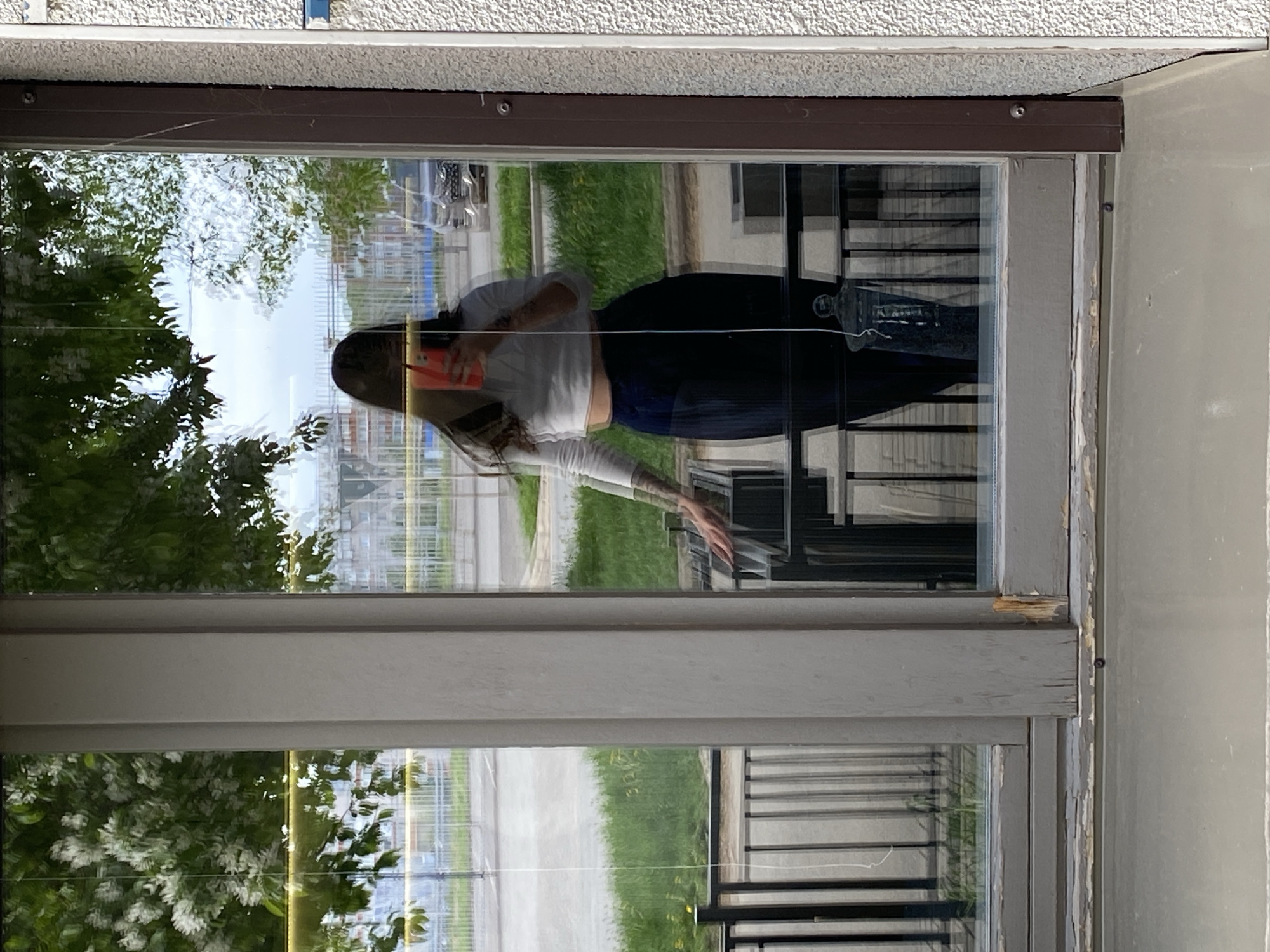 A reflection of a woman taking a selfie is visible in a window. She is holding an orange-red phone. The reflection also includes trees, green grass, and possibly a building in the background. A railing is also visible in the reflection. The window frame appears to be grey, and the exterior of the building has a textured surface. It is not possible to determine the location in the city of Votkinsk, Russia without additional information.