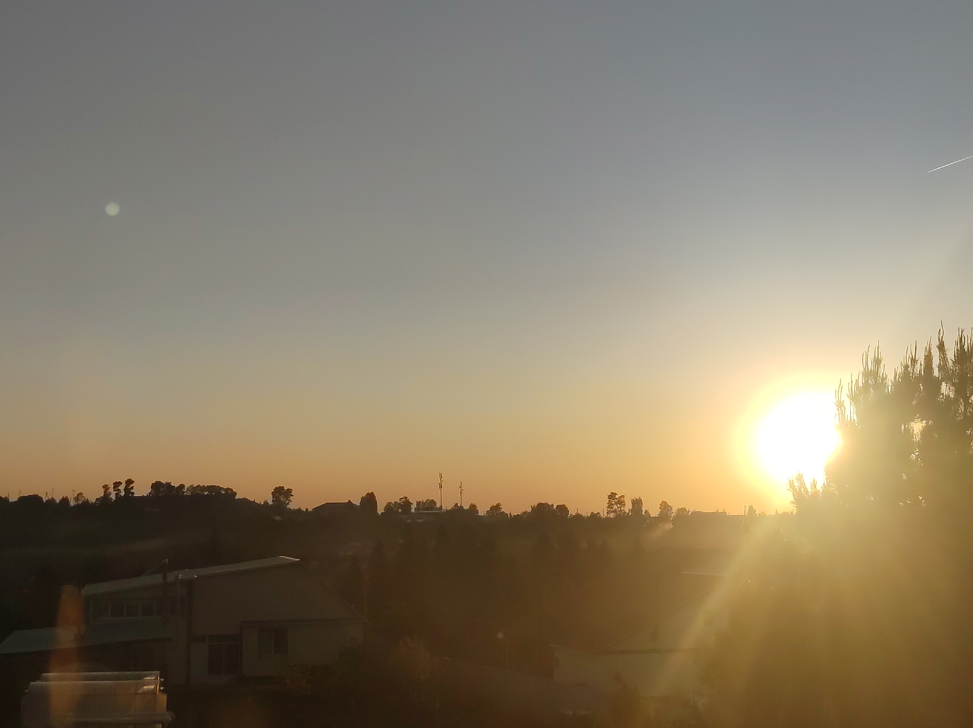 The image shows a sunrise in Фоки, Russia. The sun is a bright white-yellow disc, partially obscured by a coniferous tree on the right. The sky transitions from pale blue at the top to orange near the horizon. A contrail is visible in the upper right of the sky. A small white circular object is present in the upper left. The landscape includes silhouetted hills with scattered trees and buildings with angular roofs. A layer of haze or mist obscures the lower part of the image.