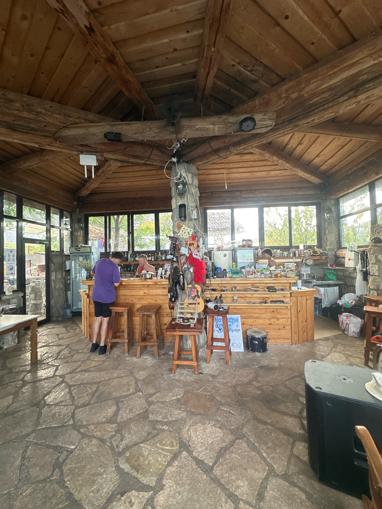 The image depicts the interior of a rustic establishment, possibly a bar or cafe, with a wooden ceiling and exposed beams. The floor is paved with irregular flagstones. In the center, a wooden counter serves as a bar area, stocked with bottles and various items. A person in a purple t-shirt and black shorts stands at the counter, facing away from the camera, seemingly interacting with the bar. Another person, with a bald head and a light-colored shirt, is visible behind the counter. The space is furnished with wooden stools. Large windows look out onto an outdoor environment with trees and buildings. There are decorative items and equipment, including what appears to be musical instruments and speakers, scattered around the space. The overall impression is of a casual, possibly open-air, gathering spot. The location is identified as Cetinje, Montenegro.