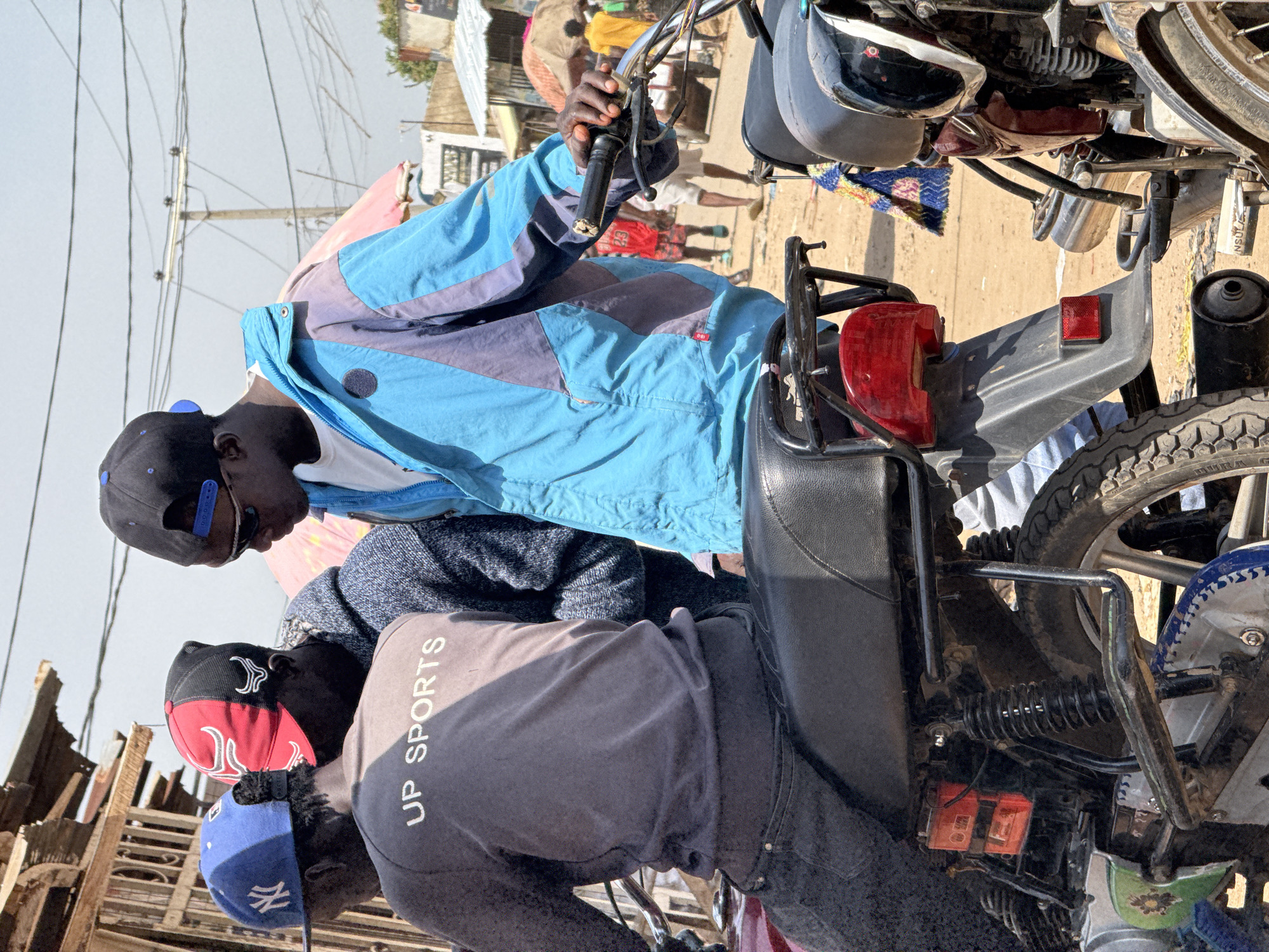 The image depicts a street scene in Abuja, Nigeria, with three young men on motorcycles. The man in the foreground on the left wears a grey long-sleeved shirt with "UP SPORTS" printed on the back, a blue New York Yankees baseball cap, and dark jeans. He is seated on a motorcycle with a black seat and a red taillight. To his right, another man, partially obscured, wears a red and black baseball cap with a bull logo and a dark hooded jacket. The man in the center is facing towards the right, wearing a bright blue and grey jacket over a white t-shirt and a black baseball cap. He is holding the handlebar of a motorcycle. In the background, there are market stalls with thatched roofs, utility poles with wires, and other people walking or standing. The ground is sandy and dusty, characteristic of an outdoor urban environment in Nigeria. The overall impression is a casual gathering of individuals with their motorcycles in a bustling street.