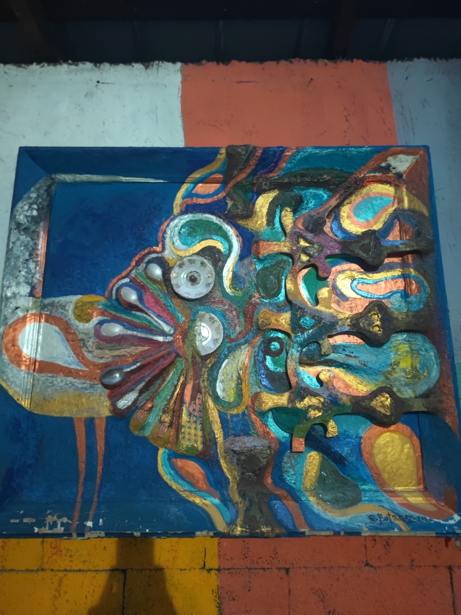 This is a photograph of a colorful, abstract relief sculpture mounted on a blue background, which is part of a larger mural or wall painting in Tuzdybastau, Kazakhstan. The sculpture itself is multi-dimensional, with various shapes and textures painted in vibrant blues, greens, golds, oranges, and reds. It incorporates found objects, including what appear to be two small ceramic dishes or saucers with floral patterns and metallic elements, possibly spoons or other metallic fragments, embedded within the painted forms. The overall style is dynamic and organic, with swirling lines and raised elements that create a sense of movement and depth.
The sculpture is framed by a textured blue border that is peeling away at the bottom, revealing lighter material beneath. The background wall above the sculpture is painted with horizontal bands of white and orange. Below the sculpture, the wall is painted with horizontal bands of yellow and orange brick patterns, with a shadow of an unseen person cast across the bottom section. The environment appears to be outdoors, given the natural lighting and the visible shadow. The context of Tuzdybastau, Kazakhstan, suggests this artwork is likely a public piece, possibly part of a street art initiative or a permanent installation in the town.