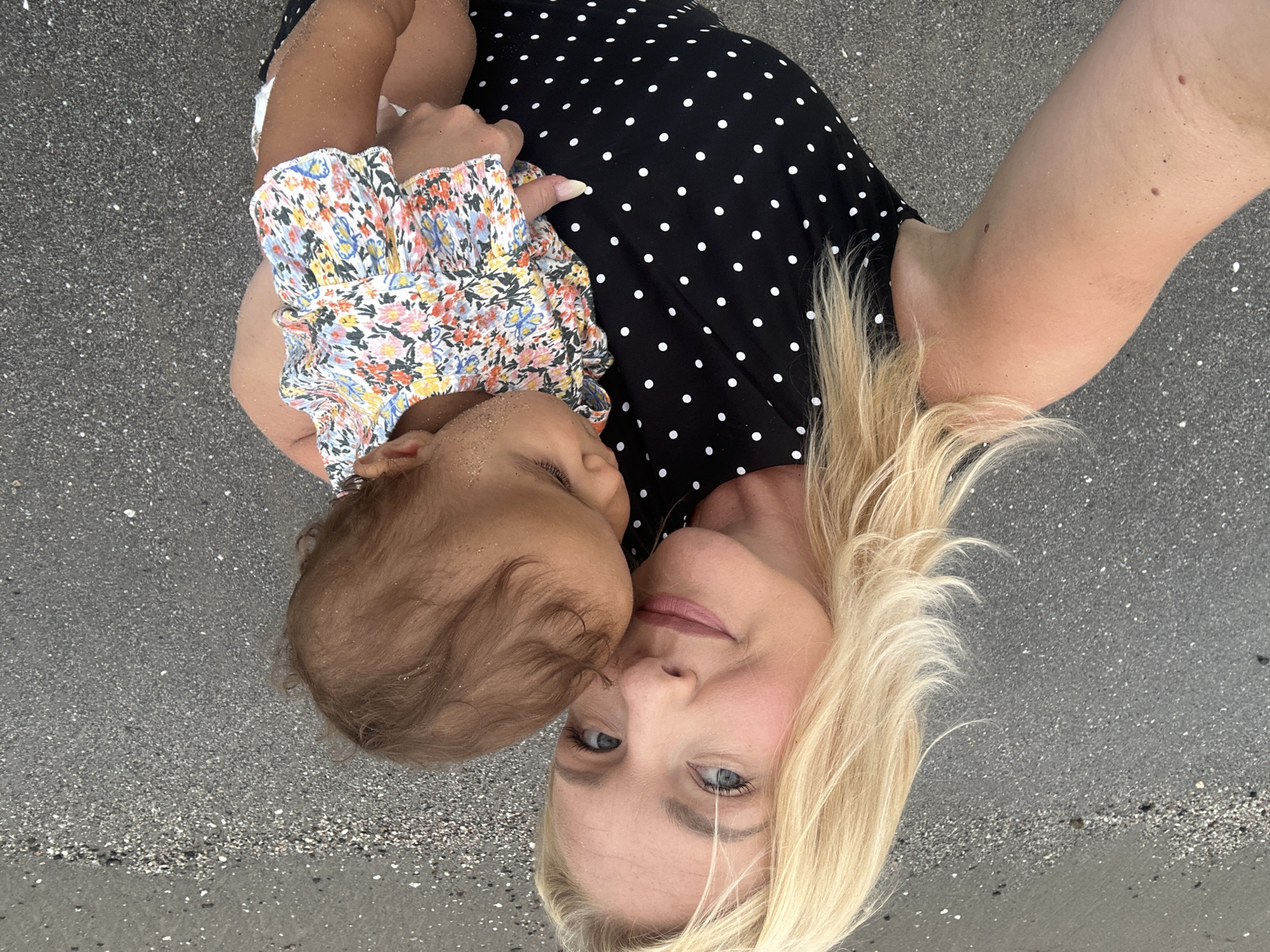 A blonde woman wearing a black and white polka dot dress is holding a baby on a beach in North Kuta, Indonesia. The woman is looking at the camera and smiling, while the baby is facing away from the camera and appears to be sleeping. The baby is wearing a floral print onesie. The beach is made of dark sand and there are small white shells scattered on it. The water is visible in the background and appears to be calm. The lighting suggests it is daytime.