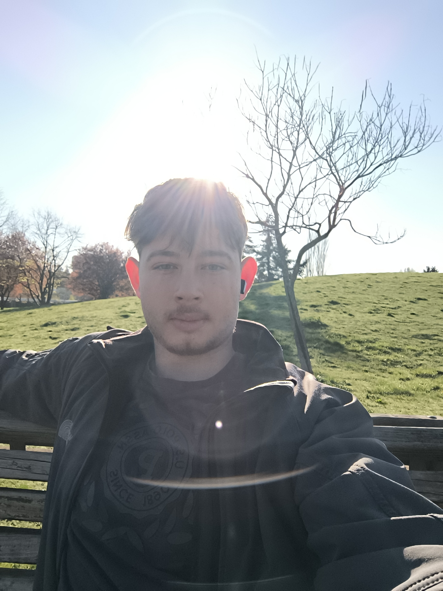 A young man is sitting on a wooden bench outdoors on a sunny day. He is wearing a dark-colored jacket over a t-shirt with a circular design. He has dark hair, fair skin, and what appears to be wireless earbuds in his ears. Behind him is a grassy hillside with trees; one tree with bare branches is prominently positioned to his right. The bright sunlight creates a lens flare and washes out parts of the image. Based on the presence of the grassy hill and the style of the trees, the location is most likely a park in the city of Sofia, Bulgaria.