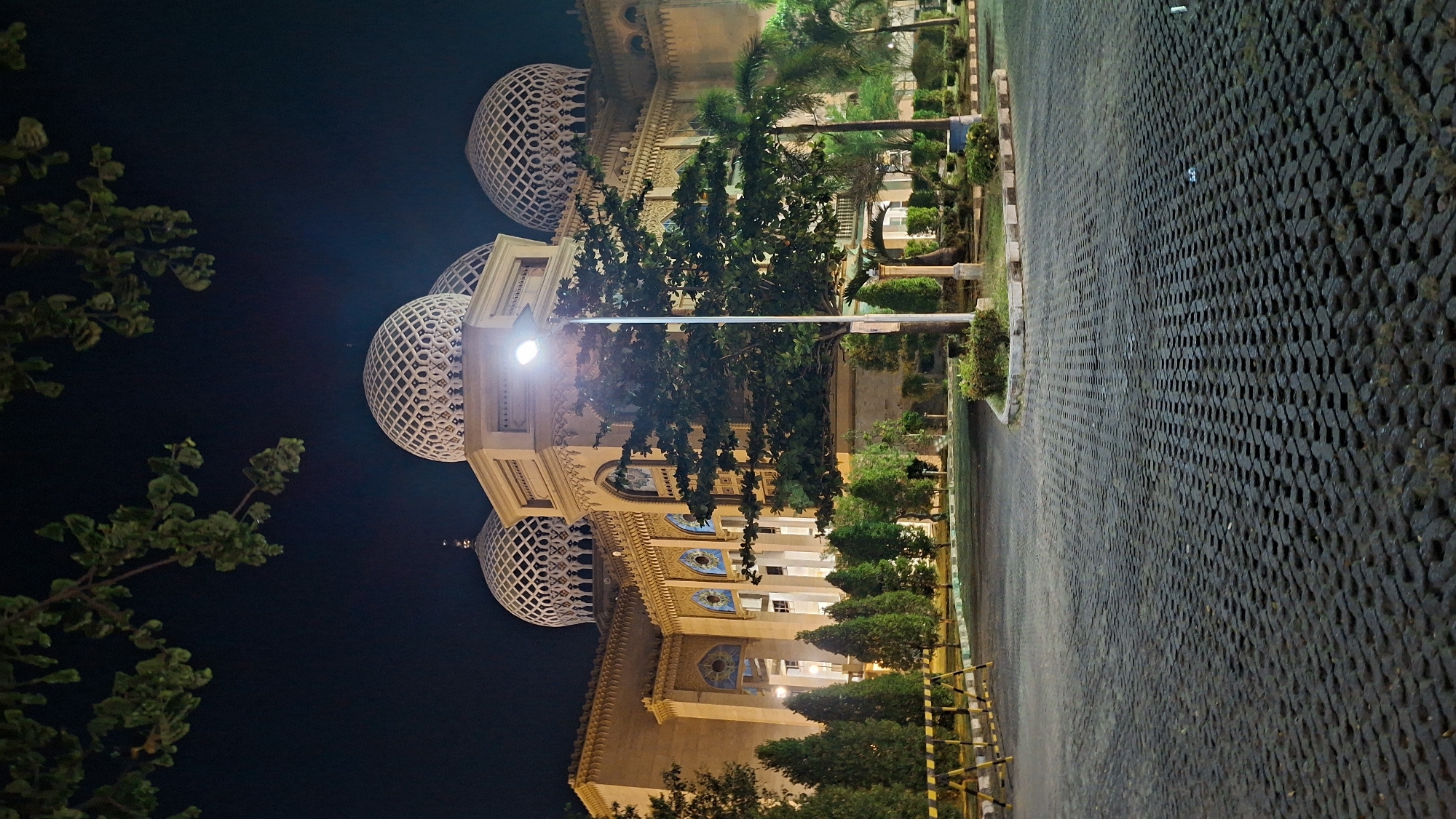 This is a night-time, low-angle, full shot of the Lhokseumawe Great Mosque (Masjid Agung Lhokseumawe) in Lhokseumawe, Indonesia. The building features multiple domes with intricate lattice work, large arched windows, and ornate architectural details. The mosque is illuminated by artificial lighting, casting a warm glow on its beige exterior. In the foreground, a paved courtyard made of interlocking concrete blocks leads towards the building. Several trees, including palm trees and smaller evergreens, are scattered around the grounds. A street lamp is visible to the right of the mosque, and safety barriers are present on the left. The sky is dark, indicating it is nighttime. No people are visible in the scene.