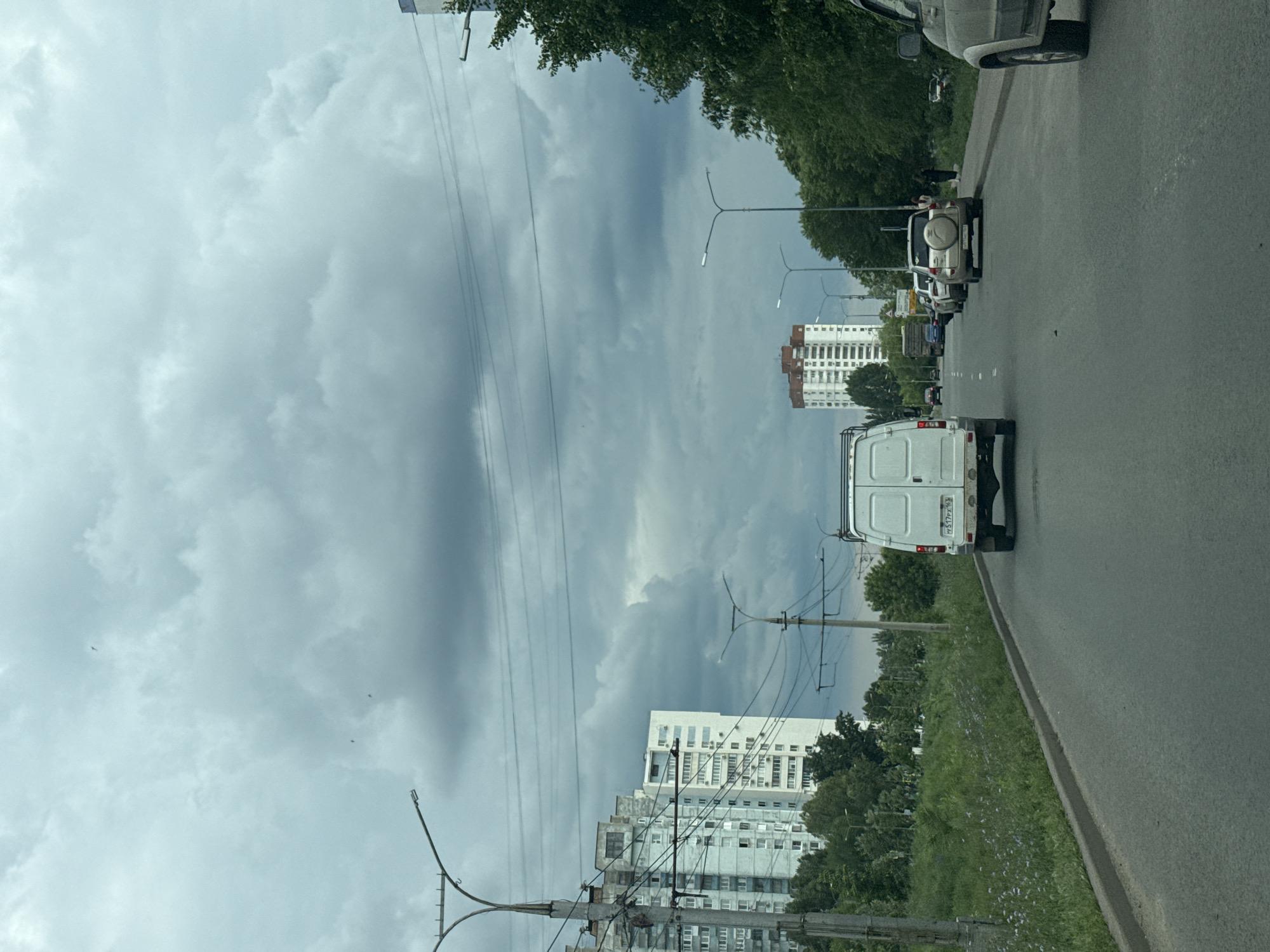 The image captures a view from a vehicle driving on a paved road, likely in a city like Samara, Russia, given the architectural style of the buildings and the presence of utility poles with extensive wiring. The sky is heavily overcast with thick, dark gray clouds, suggesting impending rain. On the left side of the road, there are tall, multi-story residential buildings with numerous windows, characteristic of Soviet-era urban architecture. A wide green embankment with grass and scattered wildflowers slopes up from the road. Utility poles with overhead power lines are prominent.
In the foreground, a white van is driving away from the camera. Further down the road, a line of parked vehicles is visible, including a light-colored SUV with a spare tire mounted on the back. On the right side of the road, there are trees and a sidewalk where two individuals are standing. The road surface appears to be asphalt, with visible lane markings. The overall atmosphere is mundane and urban, with a focus on the infrastructure and traffic flow.
