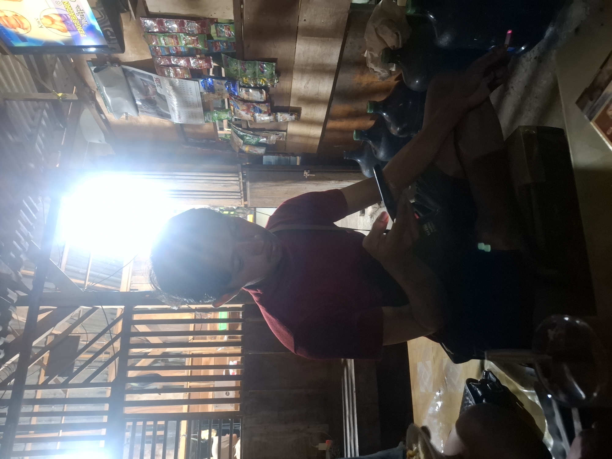 In Bulubrangsi, Indonesia, a man wearing a maroon shirt sits cross-legged on a wooden bench, looking at his phone. The environment appears to be a dimly lit, rustic establishment. Behind him, a wall is lined with shelves displaying various products, including packaged snacks and what appear to be large water jugs. A calendar is affixed to the wall, dated "2026." To the right, a television screen displays an image, although its content is unclear. Sunlight streams in from an opening in the structure, creating bright lens flares and casting shadows. The overall scene suggests a casual, everyday setting, possibly a small shop or community gathering place.