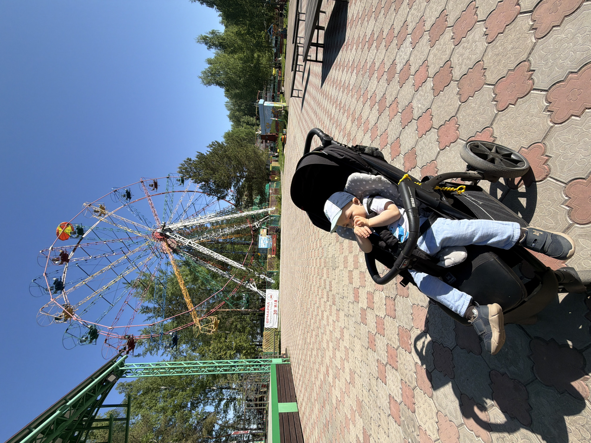 A young child is lying in a black stroller on a patterned tile surface. The child is wearing a white and blue baseball cap, a white t-shirt, and blue pants. The stroller is positioned on a patterned tile surface.
In the background, a large Ferris wheel is visible against a clear blue sky. Trees surround the Ferris wheel and other structures can be seen.
The scene is in the city of Chaikovsky, Russia.