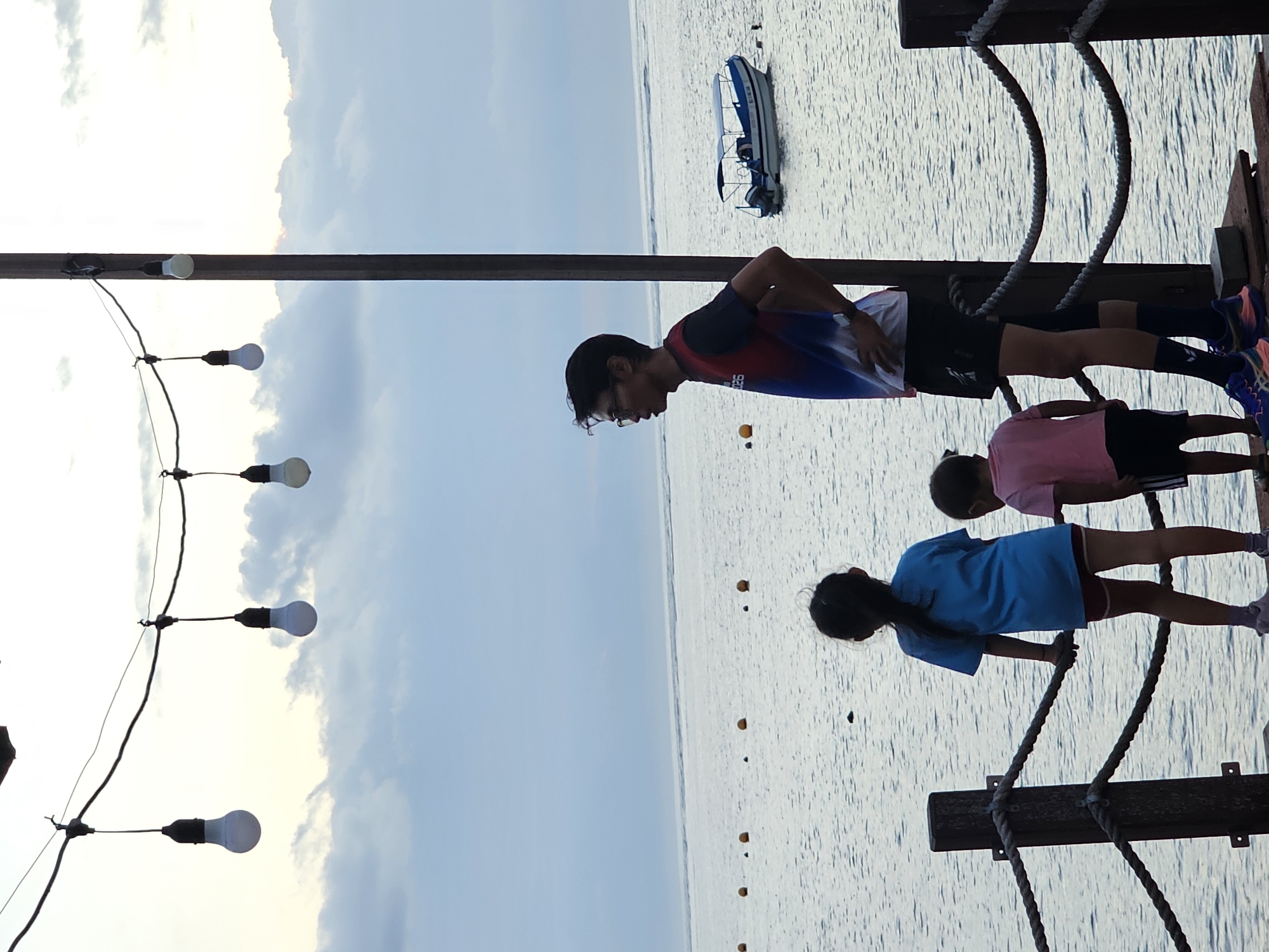 This image captures a serene moment at the beach in Denpasar, Indonesia, likely during the late afternoon or early evening given the soft light. In the foreground, a wooden deck with thick rope railings overlooks the ocean. Three people are standing on the deck, facing the water.
On the left, a young girl with dark hair in a ponytail wears a bright blue t-shirt and maroon shorts. Next to her, a smaller child, also with dark hair, is dressed in a pink t-shirt and black shorts. To the right, an adult male with glasses and short dark hair wears a colorful t-shirt with red, blue, and black accents, black shorts, and dark blue socks. He stands with one hand on his hip, observing the scene.
The ocean stretches out before them, calm and reflecting the muted tones of the sky. Several yellow buoys are visible on the water's surface, marking a section of the sea. In the middle ground, a small white and blue boat with a canopy is anchored.
Above the scene, a string of decorative light bulbs hangs from wires, casting a warm glow against the cloudy sky. The overall atmosphere is peaceful and contemplative, suggesting a family enjoying a quiet time by the sea.