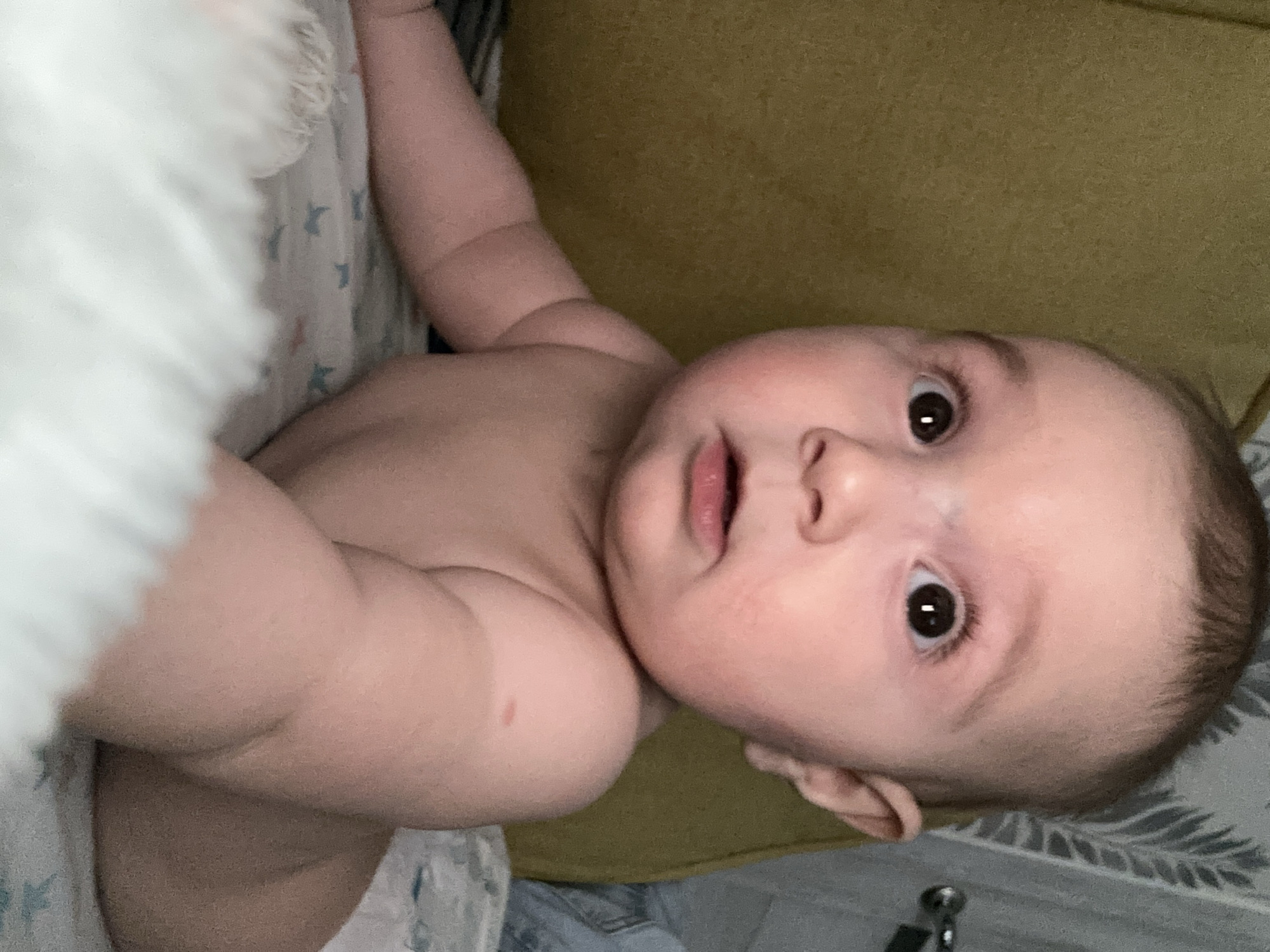 A baby is lying on their back on a surface. The baby's eyes are open, and they are looking directly at the camera. The baby is positioned between white fabric on the left and a green surface on the top. The exact location in the city of Chaikovsky, Russia, is indeterminable.