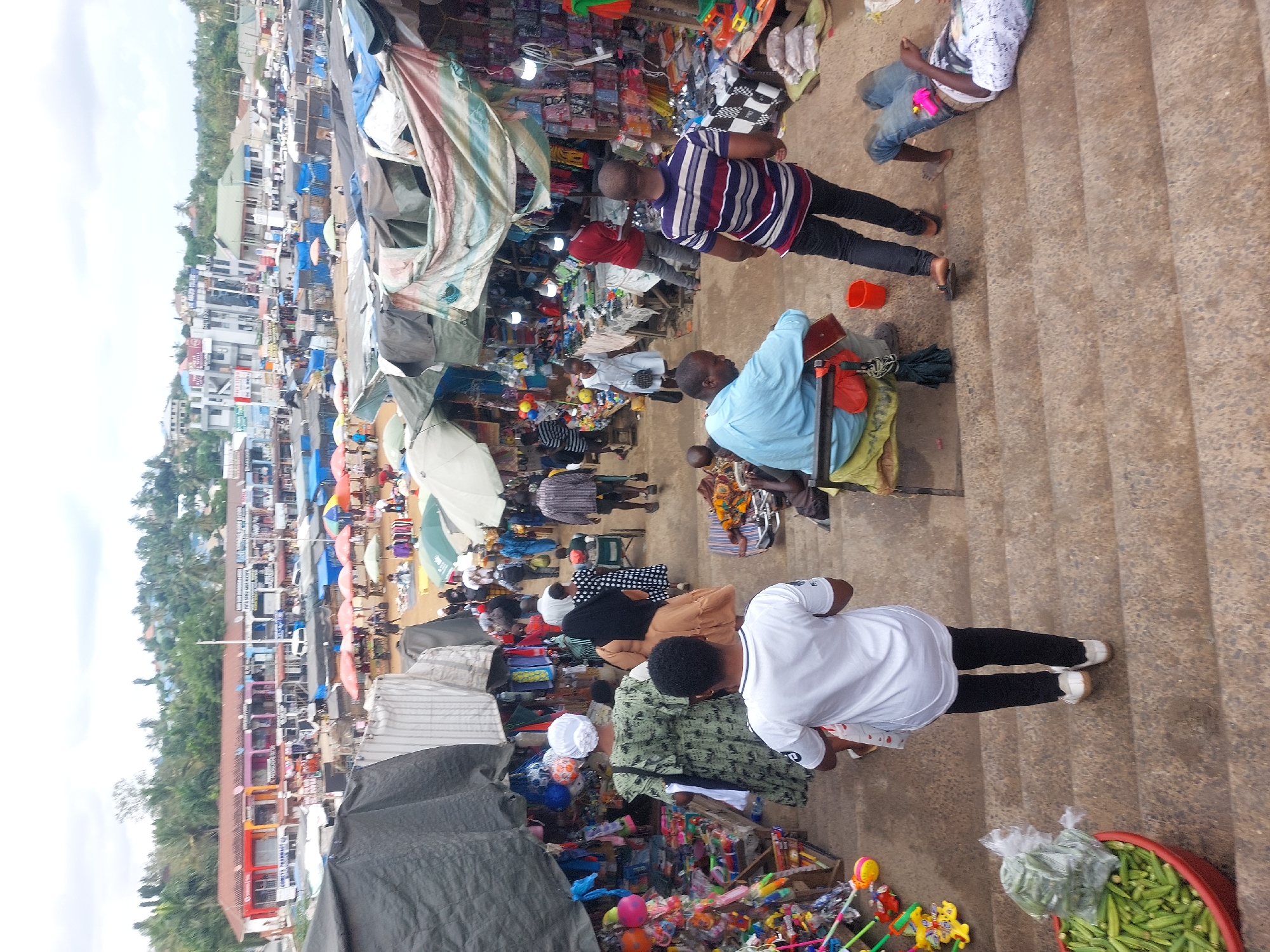 The image depicts a bustling marketplace in Dar es-Salaam, Tanzania. Vendors are selling goods under makeshift shelters and umbrellas. A variety of products are on display, including toys, okra, and other unidentified items. Several people are walking through the market, including men in striped and blue shirts, and a person in a white shirt and black pants. Some individuals are standing on a set of concrete stairs alongside the market area. The background shows buildings and trees, suggesting an urban setting.