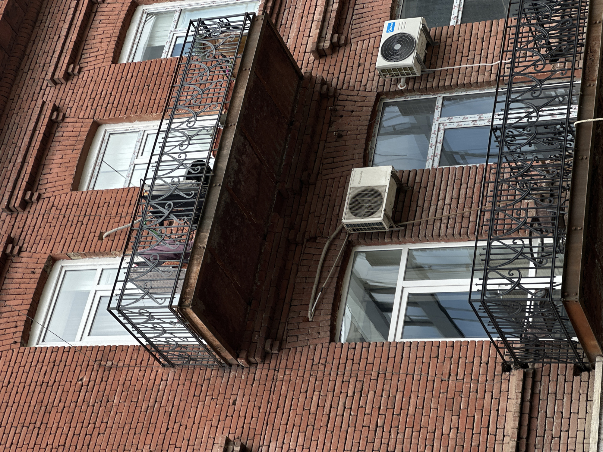 A close-up shot of a red brick building in Tbilisi, Georgia. The facade has two projecting balconies with ornate black metal railings. Windows with white frames are visible behind the balconies, and two air conditioning units are mounted on the wall between them. The brickwork is detailed, with slightly protruding sections along the vertical lines.