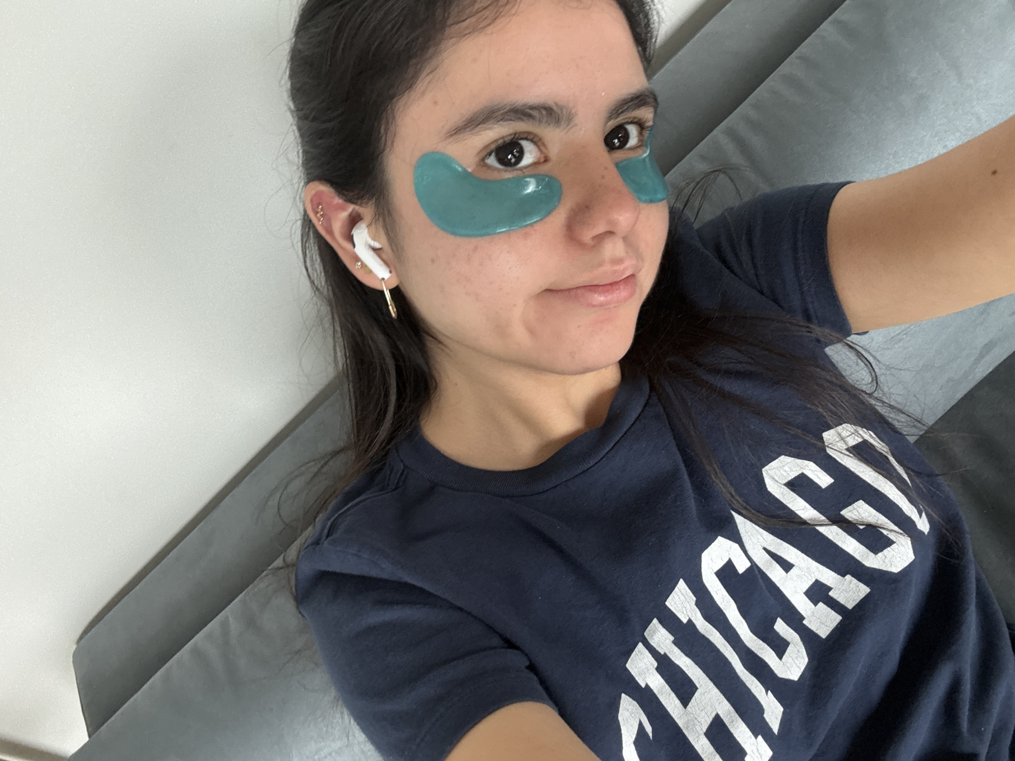 A young woman lies on a grey upholstered surface wearing a navy t-shirt with "CHICAGO" printed in white. She is wearing white wireless earbuds and has green under-eye patches on her face. The background consists of a white wall. The location cannot be determined from this image.