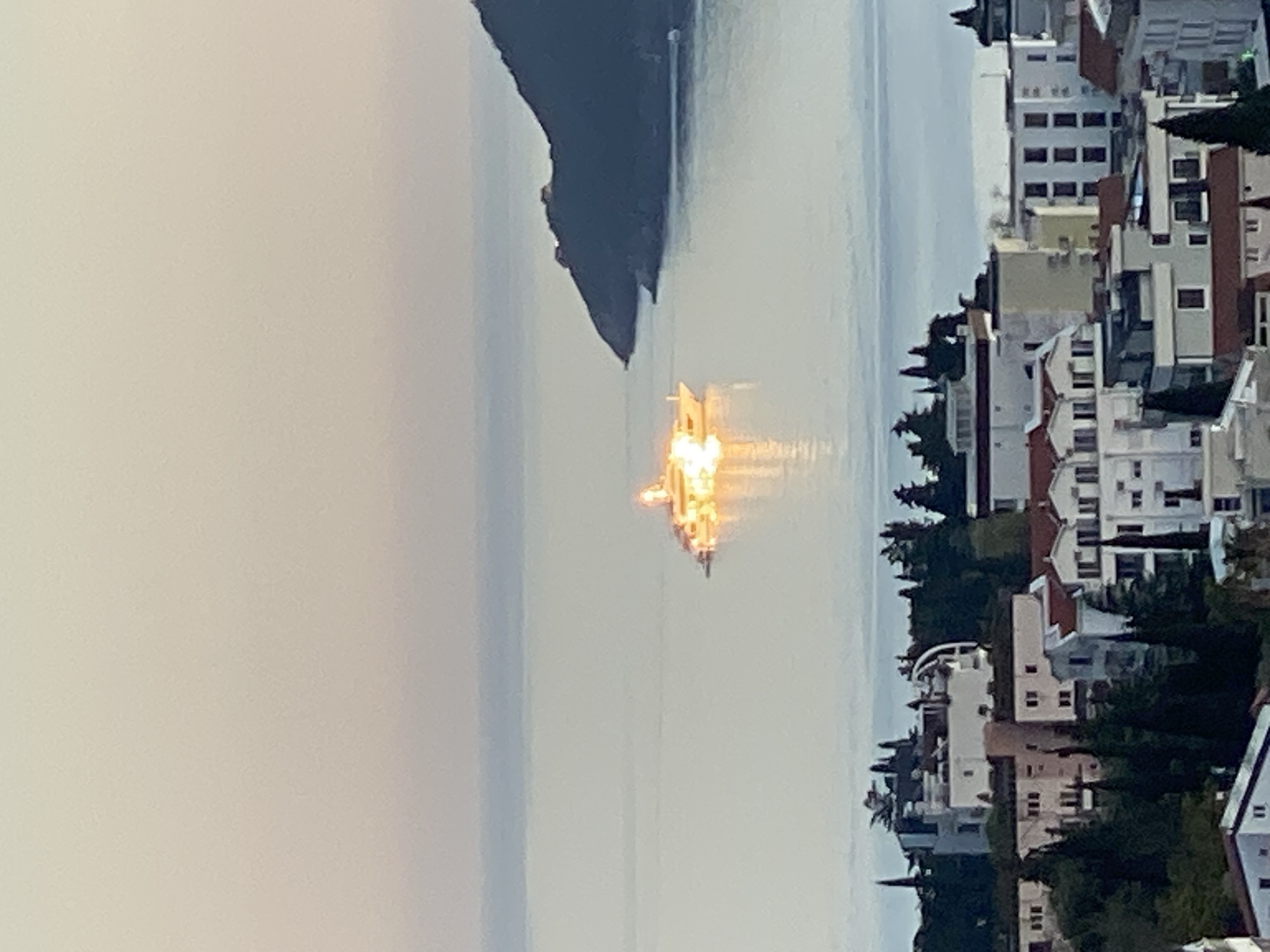 The image shows a seaside view in Budva, Montenegro. A mountainous silhouette is on the left, and houses and trees are on the right. The water in the center reflects the sunlight.