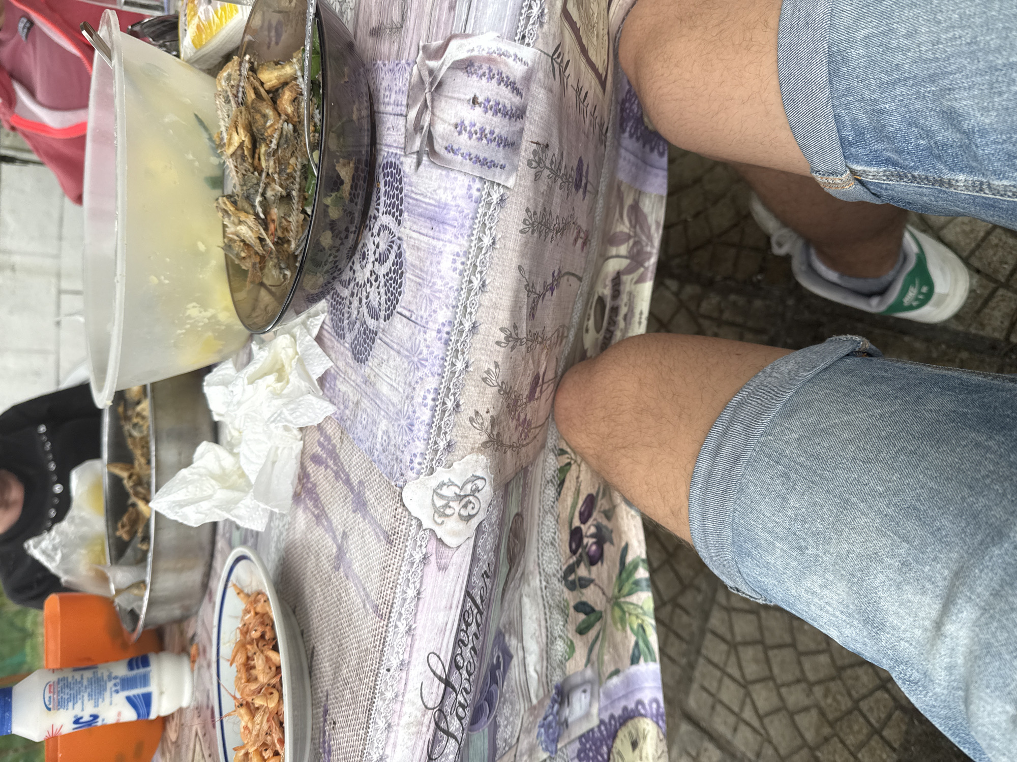 The image depicts a table laden with food. There's a large plastic container, a bowl with what appears to be fish, a plate of shrimp, and a bottle of condiments. The table is covered with a decorative tablecloth featuring a floral lavender design. A person wearing denim shorts and sneakers is sitting at the table. The setting seems to be outdoors, possibly in Zvezditsa, Bulgaria.