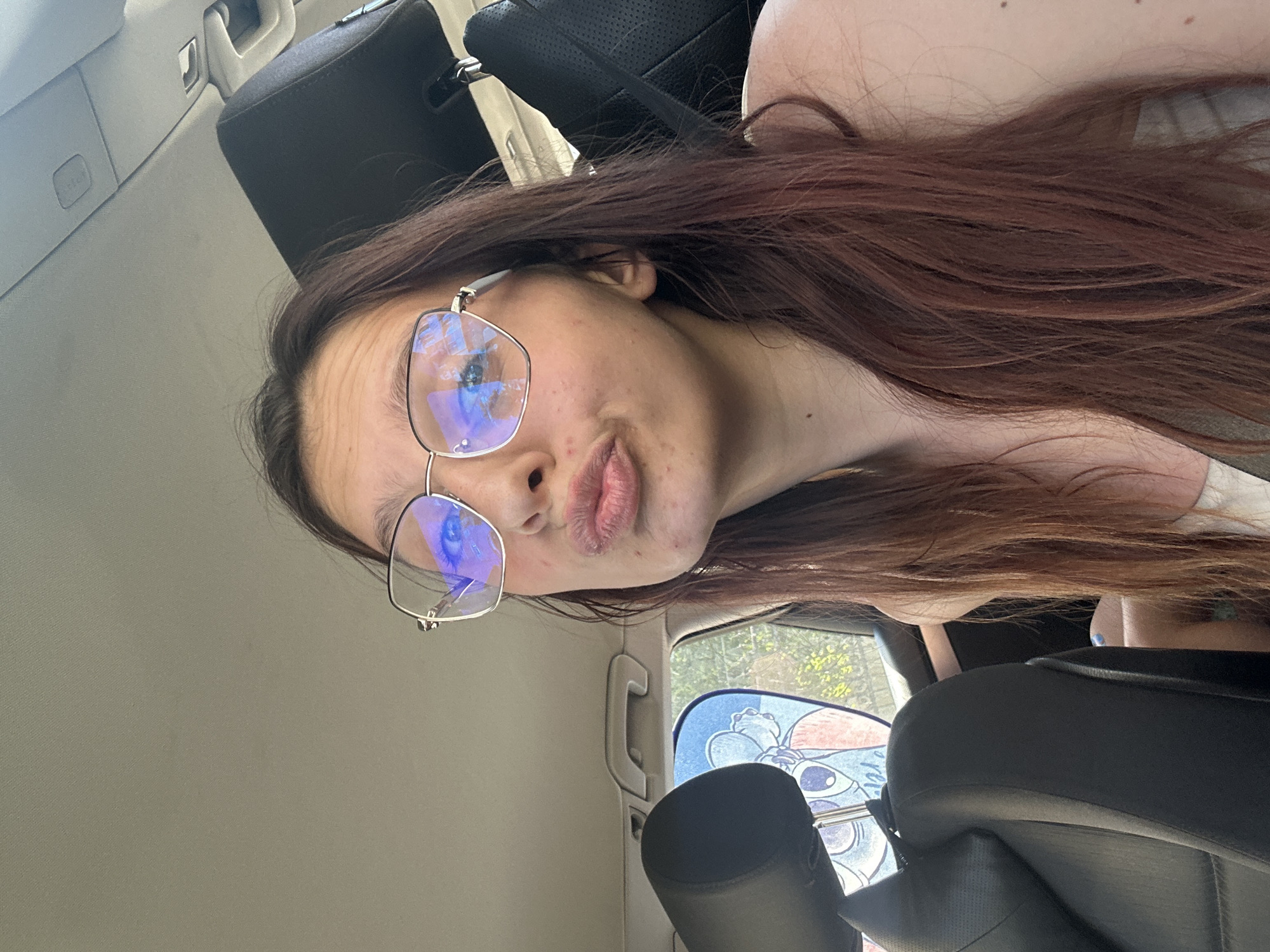 A young woman with long, dark-brown hair and glasses is in the front passenger seat of a car. She is puckering her lips and looking at the camera. The car's interior is visible, including a black headrest and a sunshade with a cartoon character on the window. The image appears to be taken during the daytime, with natural light filtering into the car. The location is not specified in the image, but it is stated to be in the city of Tryavna, Bulgaria.