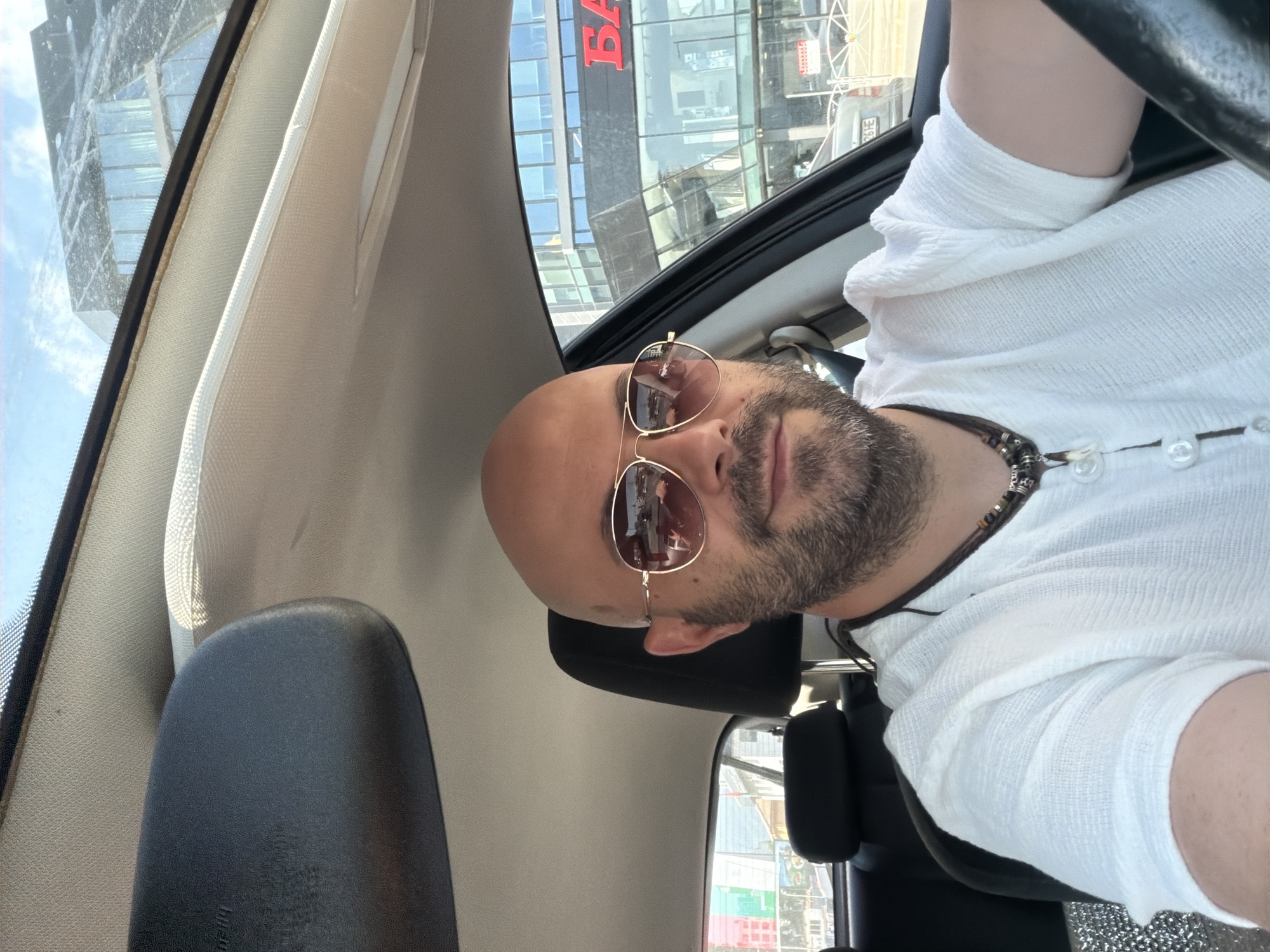 A bald, bearded man wearing sunglasses and a white shirt with a necklace is sitting in the driver's seat of a car. The car is parked in front of a modern glass-fronted building with a large red sign on it that appears to say "БАНКА" which translates to "BANK" in Bulgarian. The building is located in the city of Sofia, Bulgaria. The sky is visible through the car's windshield, and the sun is shining.