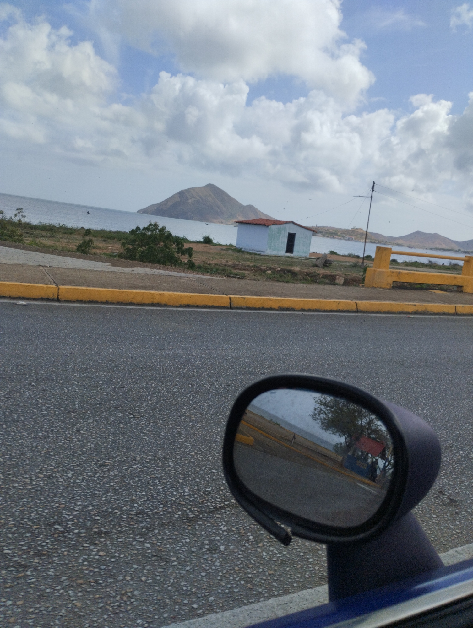 The image depicts a roadside view with a body of water and distant hills in the background. In the foreground, a section of a road is visible, with a yellow curb along the side. A car's side mirror is prominently featured, reflecting a scene that includes trees and what appears to be a small market stall with people and a red canopy. In the middle ground, there is a small, light-blue building with a reddish roof and a dark doorway, situated on a grassy area near the water. The sky is partly cloudy, with white clouds against a light blue background. The overall scene suggests a coastal environment, possibly in or near a town or village. The reference to Juan Griego, Venezuela, places the scene in a specific geographical context, implying a coastal town known for its bay and historical significance.