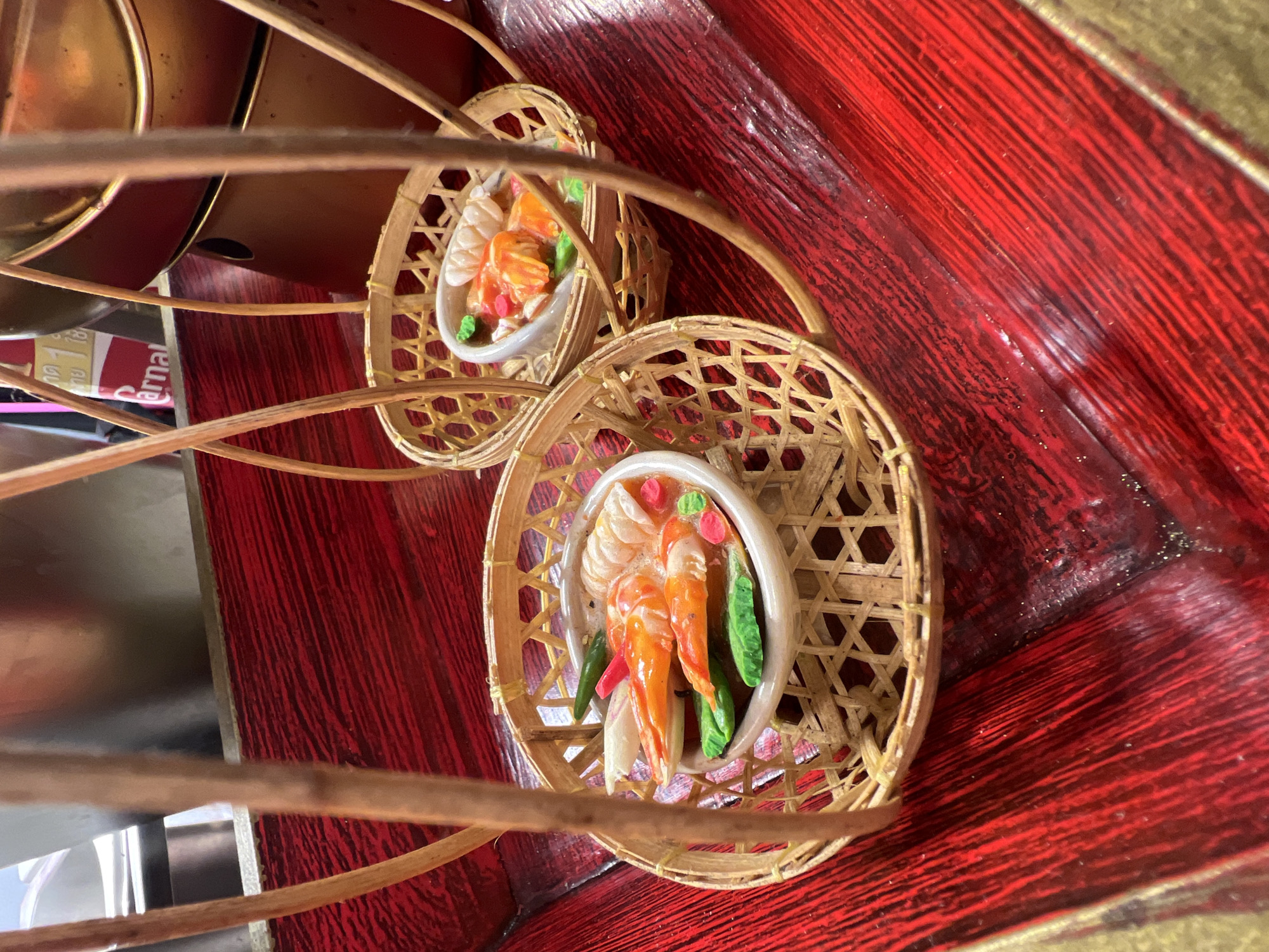 The image displays two miniature food replicas in woven bamboo baskets, presented on a textured red surface. The replicas appear to be of seafood dishes, with prominently featured orange shrimp in a broth, garnished with green and pink elements. The baskets are supported by light-colored wooden poles, suggesting a display or decorative arrangement. In the background, metallic objects and a partially visible sign with Thai text are present, indicating a possible restaurant or market setting in Karon, Thailand. The red surface exhibits a distressed, striated texture, possibly representing wood or a similar material.
