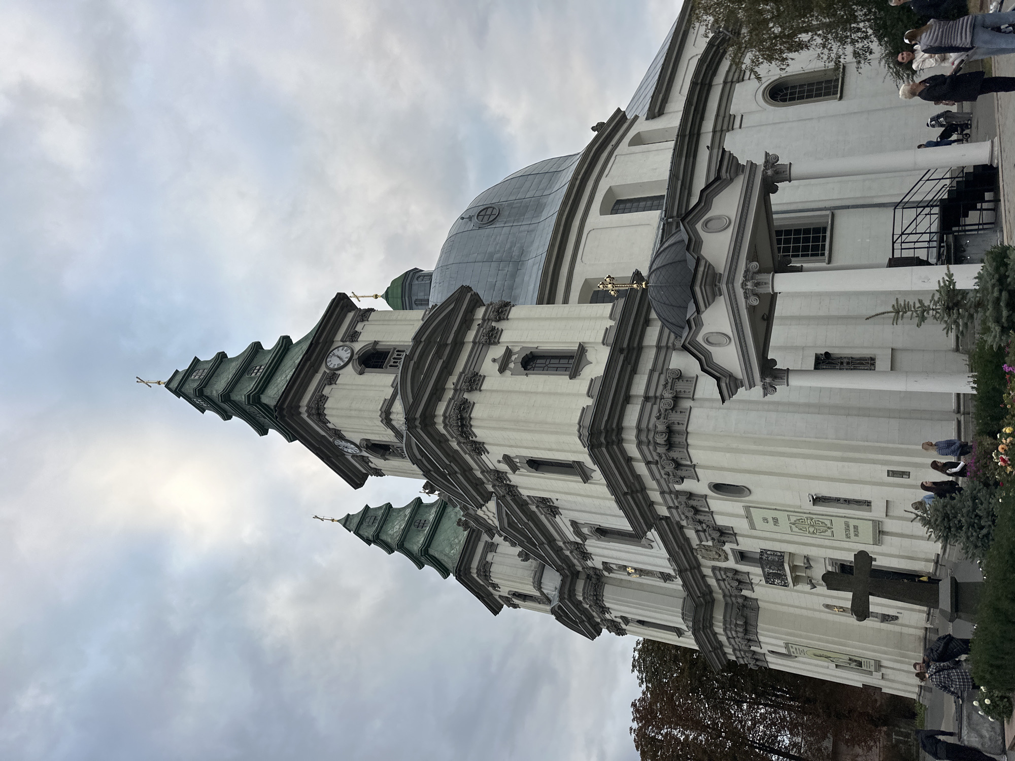 The image shows a low-angle, full shot of a large, ornate church in Ternopil, Ukraine, under a cloudy sky. The church is a white Baroque-style building with gray architectural detailing. It features multiple green-roofed spires topped with crosses, a large central dome, and decorative facades with columns and window frames. A prominent, dark stone cross stands in the foreground to the left of the church entrance. Several people are visible in front of the church, some standing near the cross and others walking towards or away from the building. The environment appears to be an urban setting, with trees visible to the left and right of the church, suggesting it is part of a city square or park. The lighting is overcast, casting a diffused light on the scene. The overall impression is one of grandeur and religious significance.
