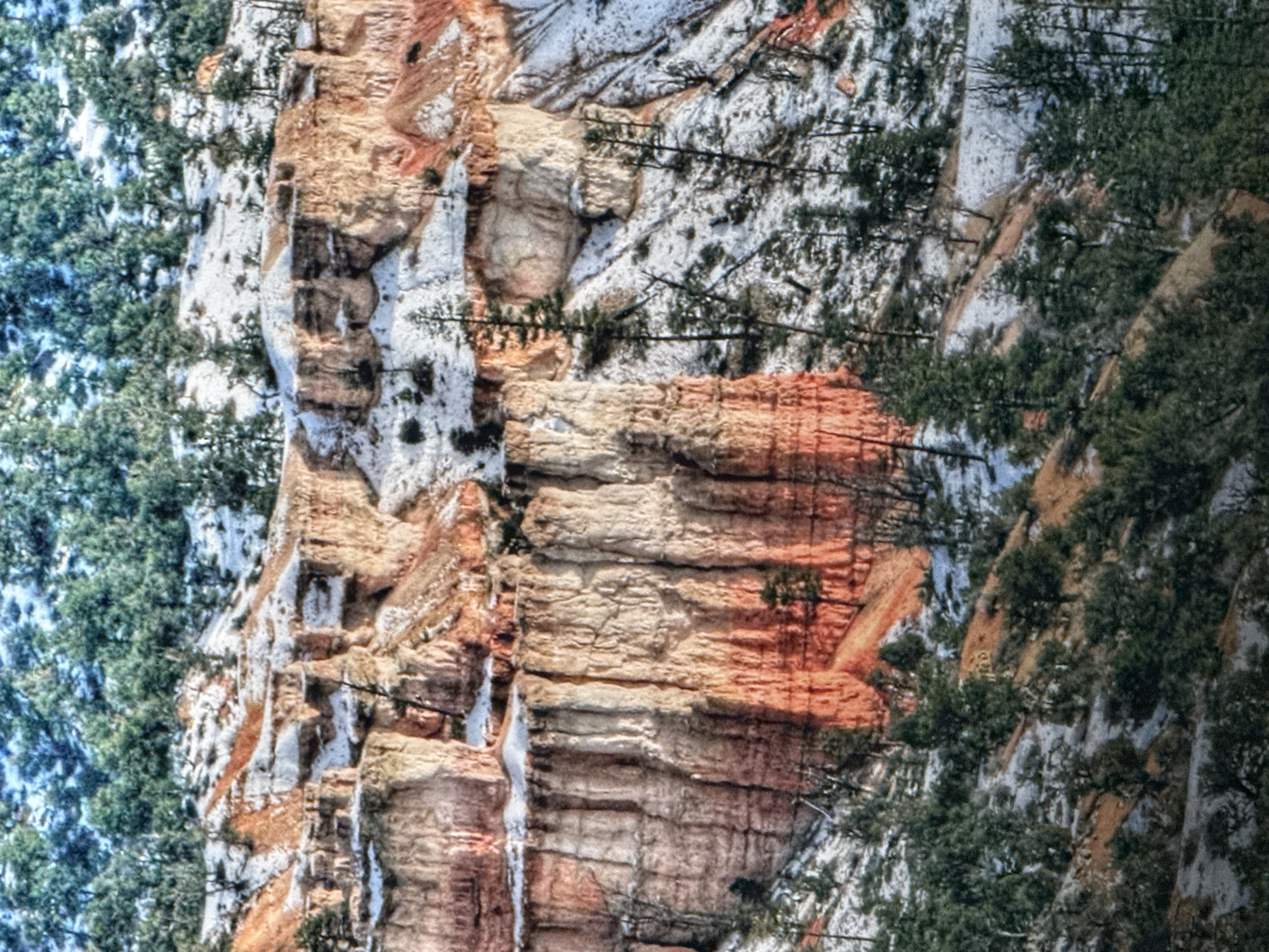 The image depicts a snow-dusted landscape with towering, striated rock formations, likely hoodoos, characteristic of Bryce Canyon National Park in Utah. The rocks display varying shades of orange, pink, and white, with patches of snow highlighting their contours. Evergreen trees are scattered across the scene, some clinging to the slopes and others sparsely dotting the tops of the formations. The ground is covered in a thin layer of snow, with areas of exposed reddish-brown earth peeking through. The overall impression is one of a rugged, natural environment, possibly in late autumn or early winter. The image does not show any people, city elements, or indications of Saratov, Russia.