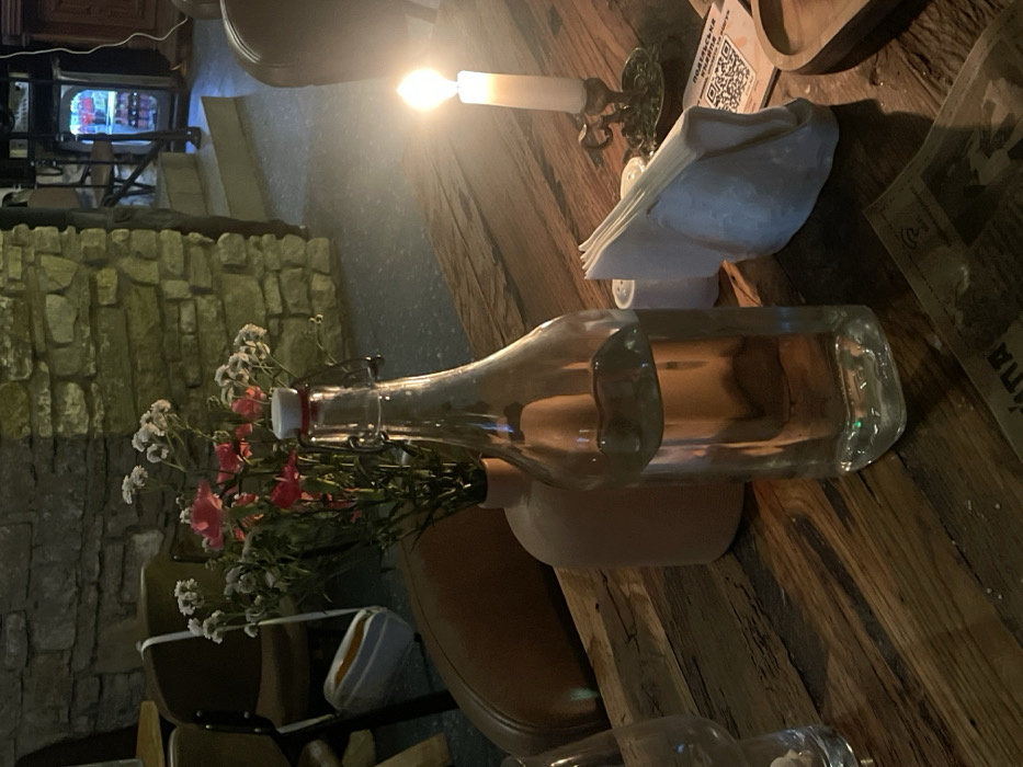 A clear glass bottle with a hinged lid rests on a light brown vase, holding a small bouquet of white and pink flowers. The bottle and vase sit on a wooden table. A candle in a metal holder casts a warm glow, next to a stack of white napkins. A stone wall is visible in the background, and portions of chairs are in the lower left.