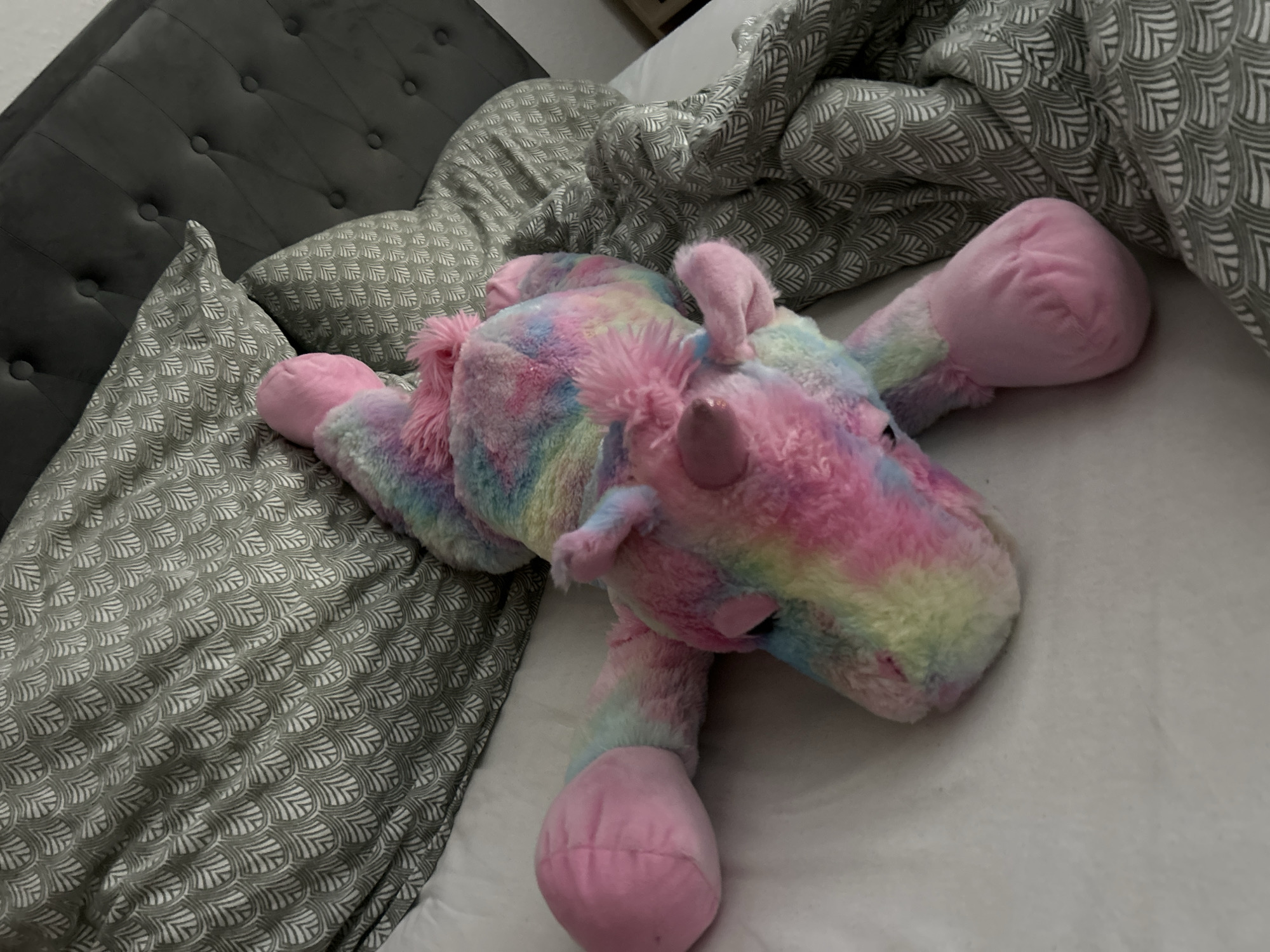 A stuffed unicorn toy with rainbow-colored fur lies on a bed. The unicorn is positioned on its side, with its head facing right and its legs splayed out. The bed has a gray tufted headboard and is covered with pillows and a comforter, both of which feature a repeating gray and white scale-like pattern. The scene is indoors.