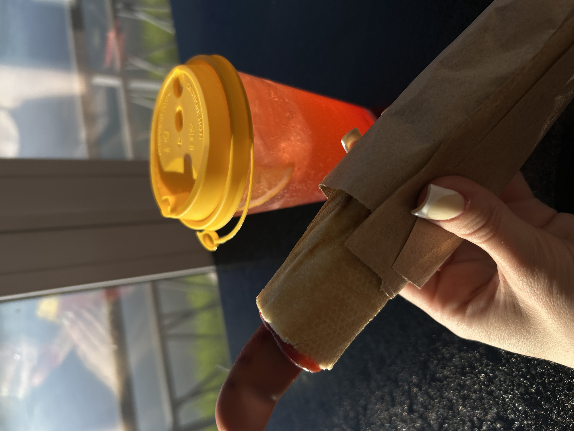 The image presents a close-up, angled view of a person holding a hot dog, with a drink visible in the background. A fair-skinned hand, featuring manicured nails with a light two-toned design, grips a hot dog partially unwrapped from a brown paper sleeve. The hot dog has a reddish sausage protruding from a light-colored bun, with a trace of red condiment, likely ketchup, visible on the sausage.
Behind the hand and food, a translucent orange or red cup with a bright yellow lid is prominent. The lid clearly displays the English warning "CAUTION HOT!". Below this text, characters resembling Asian script (possibly Chinese "注意" for "caution") are also visible. The liquid inside the cup appears light orange and contains small bubbles, suggesting a carbonated beverage.
The objects rest on a dark, speckled surface, possibly a table or counter. The background is blurred but reveals bright reflections on a window, indicating an outdoor scene with strong sunlight. Hints of greenery, sky, and indistinct shapes, possibly structures or figures, are reflected in the glass. The vibrant lighting, sharp shadows, and reflections suggest it is daytime, likely morning or afternoon. The scene captures a casual moment of enjoying a snack or meal.