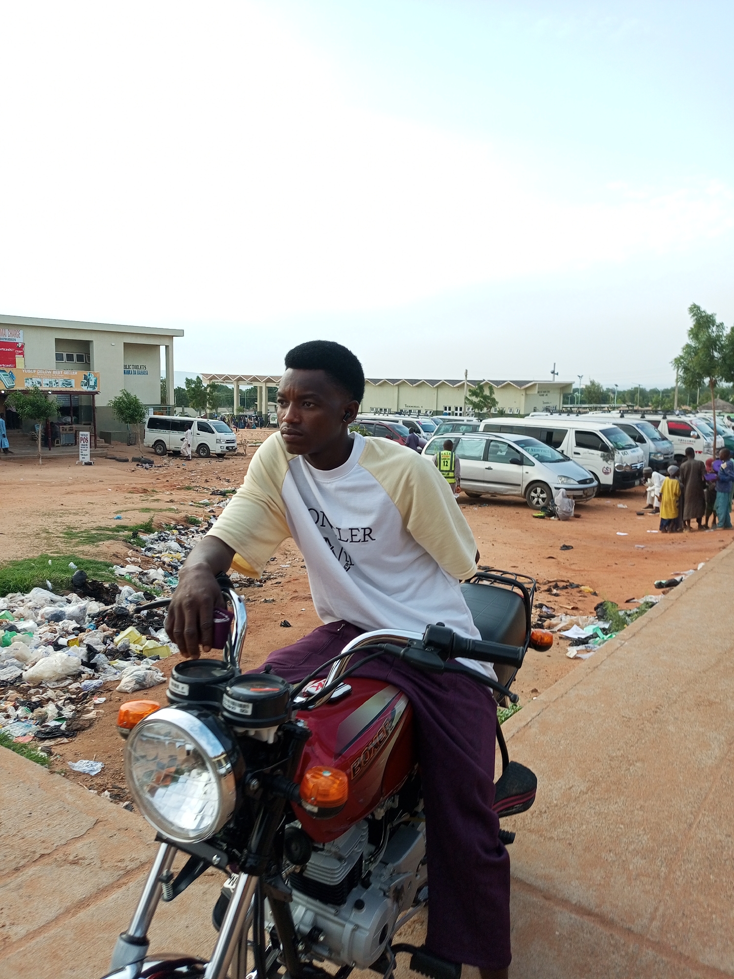The image captures a scene in Gombe, Nigeria, likely near a transportation hub or a public area with scattered refuse. In the foreground, a young man is seated on a red motorcycle. He wears a white t-shirt with yellow sleeves and dark purple trousers. His gaze is directed to the right, away from the camera. The motorcycle is the central object in the immediate foreground, with its headlight and handlebars clearly visible.
Behind the motorcycle, the ground is covered in a significant amount of scattered trash, including plastic bags and other debris, indicating a lack of waste management. In the mid-ground, several vehicles are parked in rows, including vans and cars, suggesting a parking lot or a terminal area. Some people are visible standing around these vehicles, engaged in various activities, with some appearing to be interacting.
Further in the background, a long, light-colored building with multiple windows is visible, which could be a terminal building or a commercial establishment. The sky is overcast, suggesting a cloudy day. The overall environment appears to be an urban or semi-urban setting, characterized by a mix of infrastructure and visible waste.