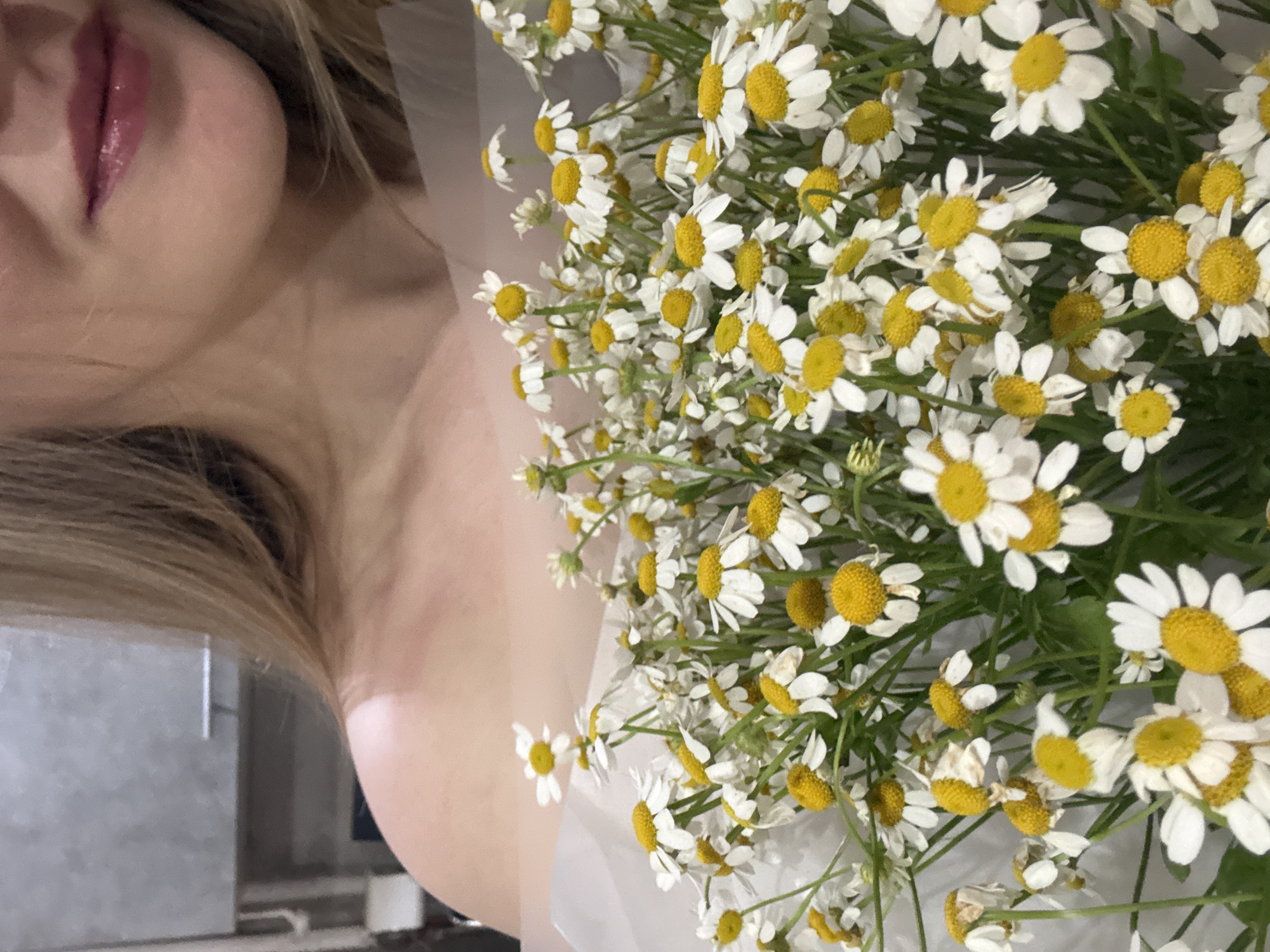 The image features a woman with blonde hair and pink lipstick holding a bouquet of chamomile flowers. Her shoulder and neck are visible. The background is blurred. The location is not discernible from the image.