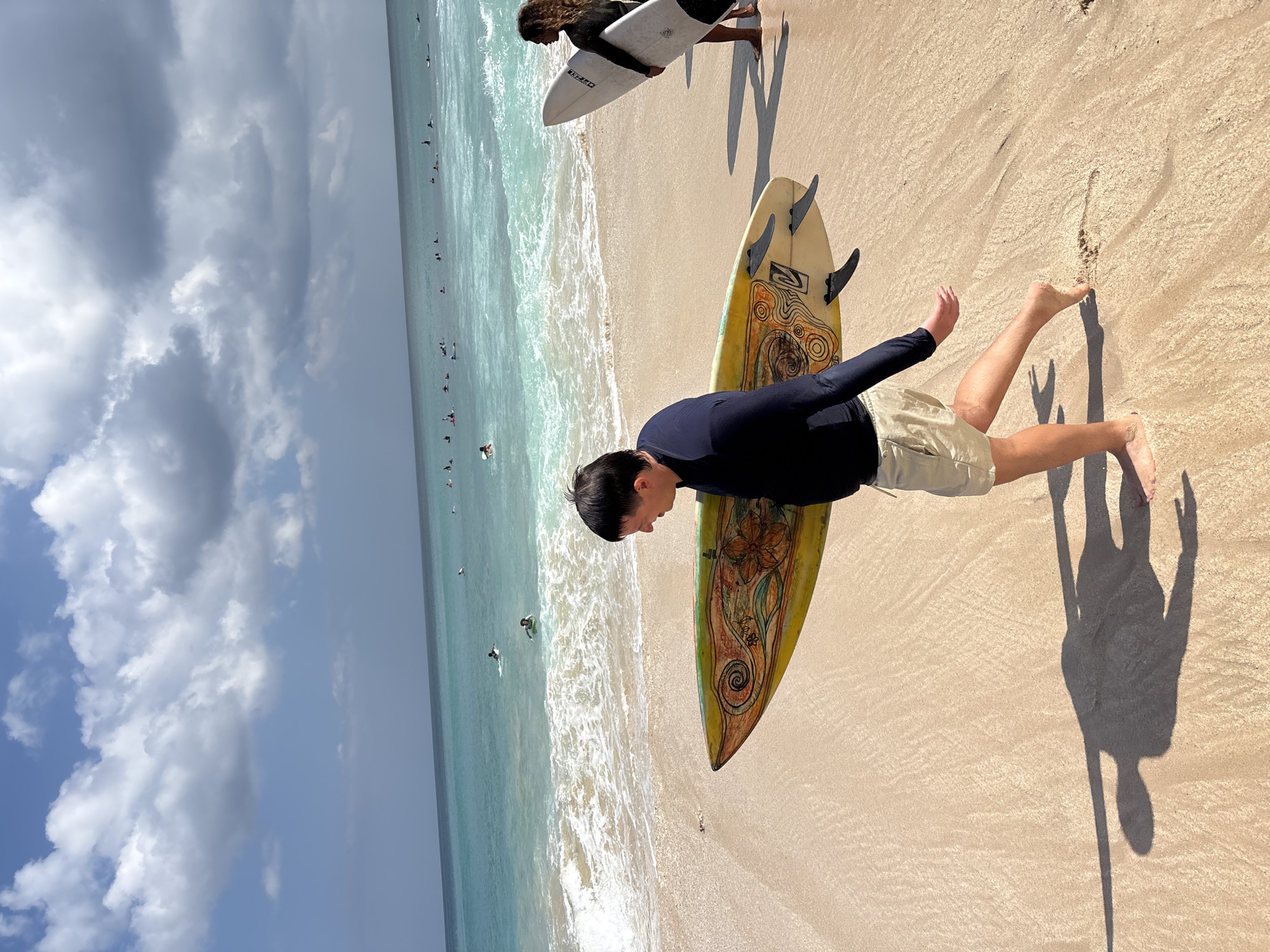 In Kuta Selatan, Indonesia, a man with dark hair and a black long-sleeved rashguard walks barefoot on a sandy beach, carrying a yellow surfboard with an orange and green decorative design. He wears beige shorts. Behind him, to the right, another person with long curly hair and a black rashguard stands holding a white surfboard. The ocean, with turquoise water and white waves, is visible in the background, populated by numerous surfers in the water. The sky is blue with scattered white clouds. The scene captures a typical day at a popular surfing spot.