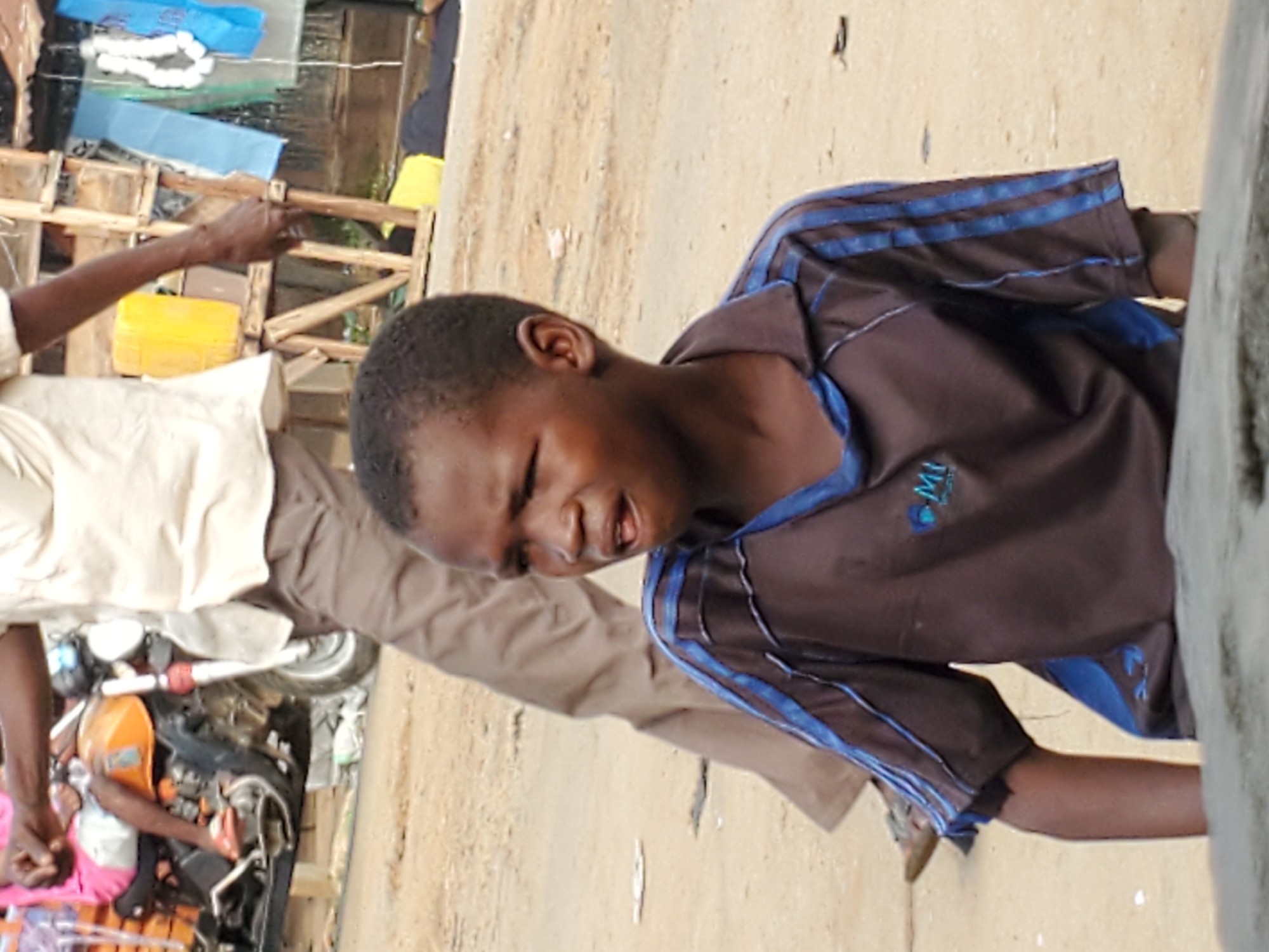 The image captures a scene in Mararaba, Nigeria. A young boy wearing a brown and blue shirt with visible logo is partially visible on the right. Another person, wearing a tan shirt and pants, is seen on the left with a hand reaching towards a wooden structure. In the background, a motorcycle lies on its side. The environment appears to be an outdoor, possibly roadside setting.