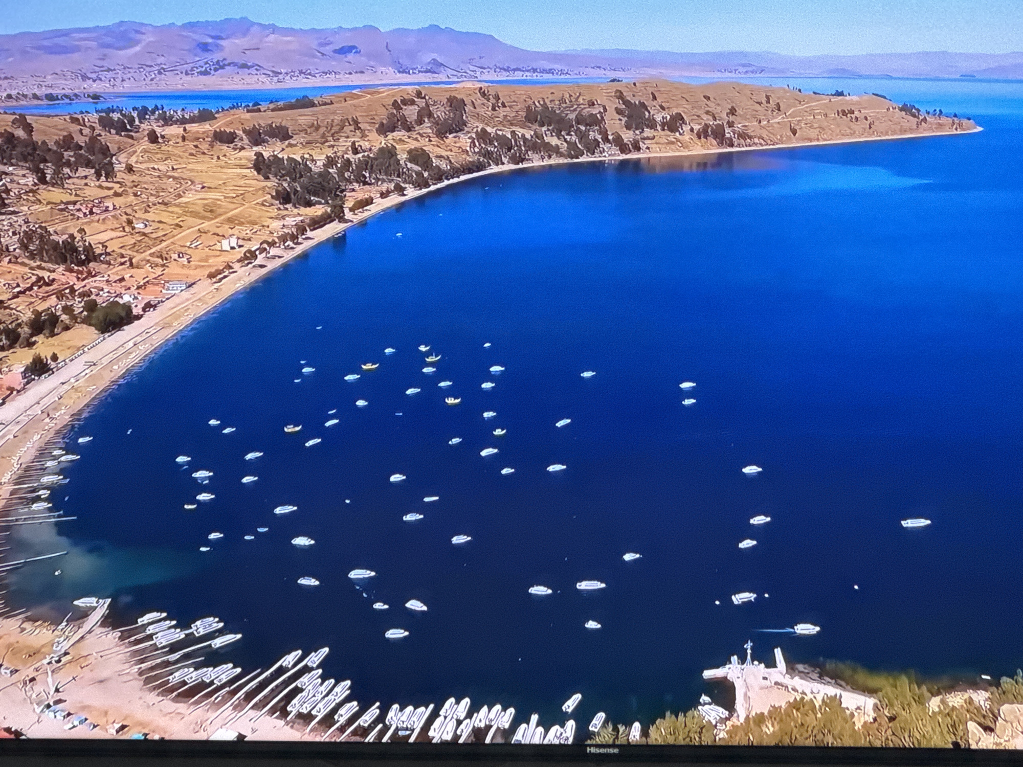 This aerial view captures a vibrant scene on a large, deep blue body of water, likely a lake. The shoreline is lined with a sandy beach and a settlement with numerous buildings and trees. A prominent feature is the vast number of small boats, mostly white, dotting the water, with a particularly dense concentration near the shore. The landscape beyond the settlement is hilly and arid, with some vegetation, and in the distance, hazy mountains are visible under a clear, bright sky. The image does not depict the city of Ləcət, Azerbaijan.