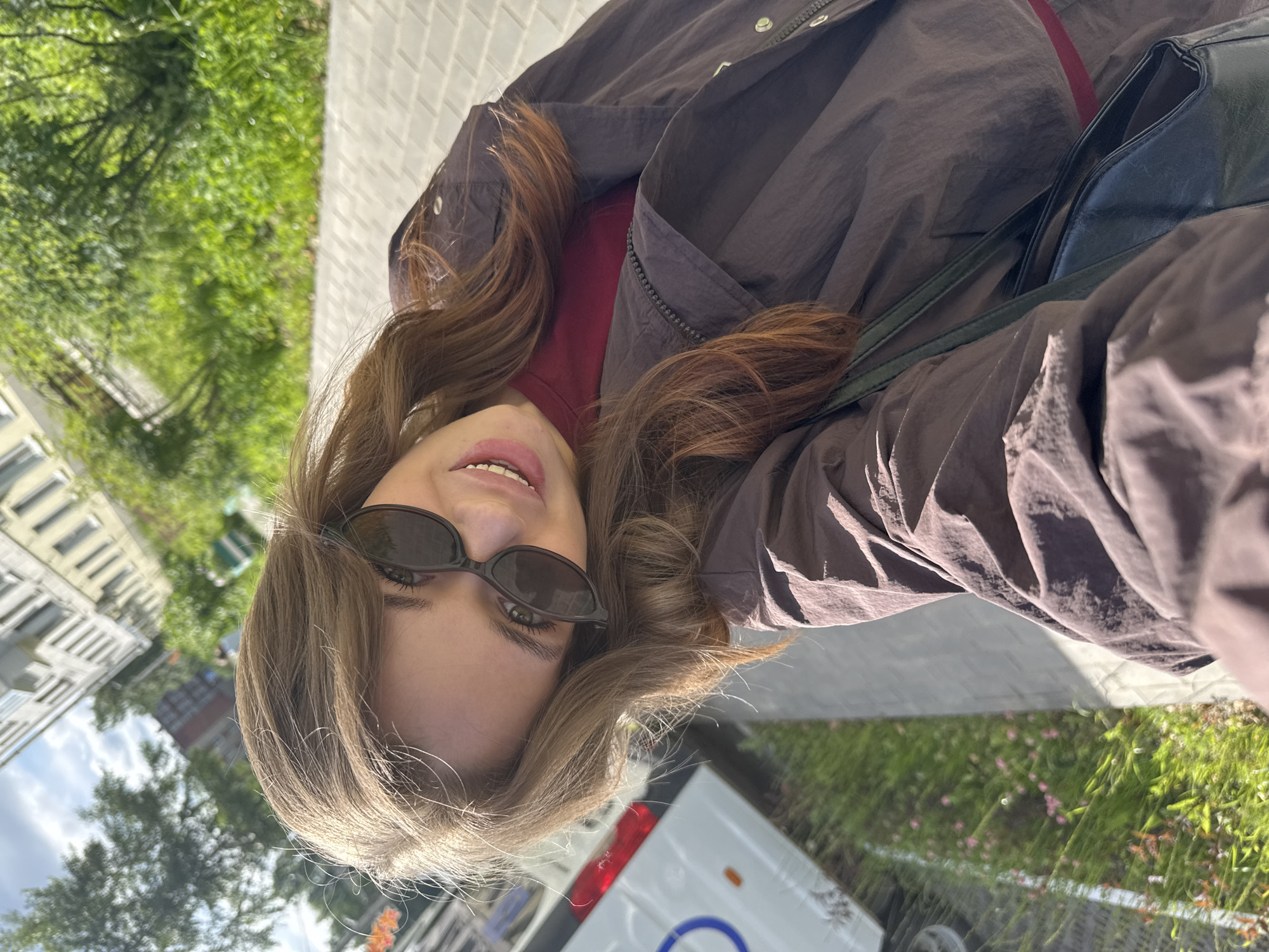 A young woman with long, light brown hair and dark sunglasses is taking a selfie outdoors on a sidewalk. She is wearing a dark brown jacket over a maroon shirt. A black purse is slung over her shoulder. In the background, there is lush green foliage, a light-colored building, and a white vehicle. The setting appears to be a city street in Chaikovsky, Russia, given the context of the request. The ground is paved with light gray rectangular tiles. The lighting suggests it is daytime.