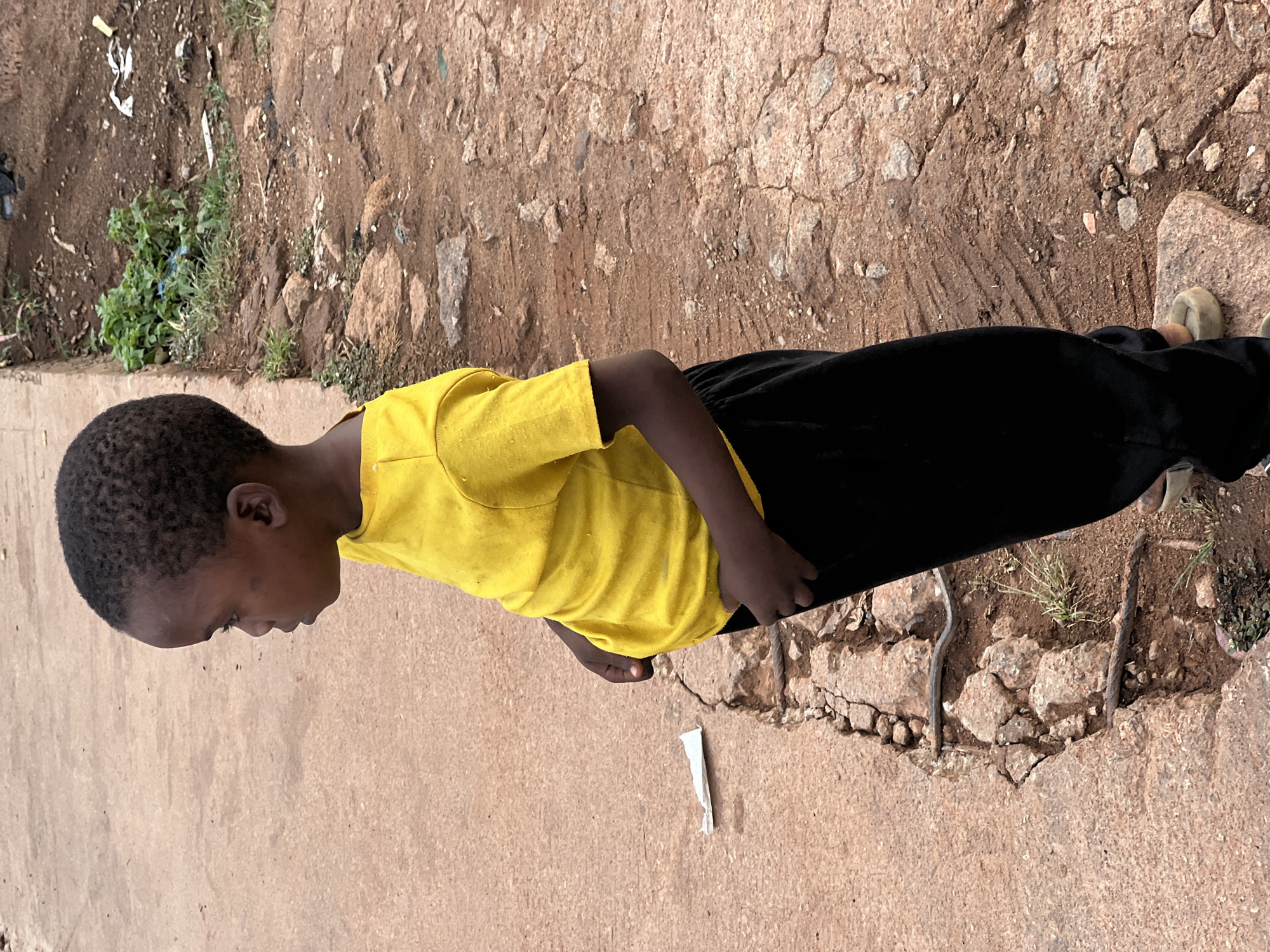A young person wearing a yellow t-shirt and black pants stands on a cracked concrete surface in what appears to be Gura Top, Nigeria. To the right of the individual is a more eroded area of ground, with visible rocks and exposed metal reinforcing bars. A piece of white trash is near the person's left foot. The background shows some vegetation growing in the cracked area. The photograph is taken from a high angle.