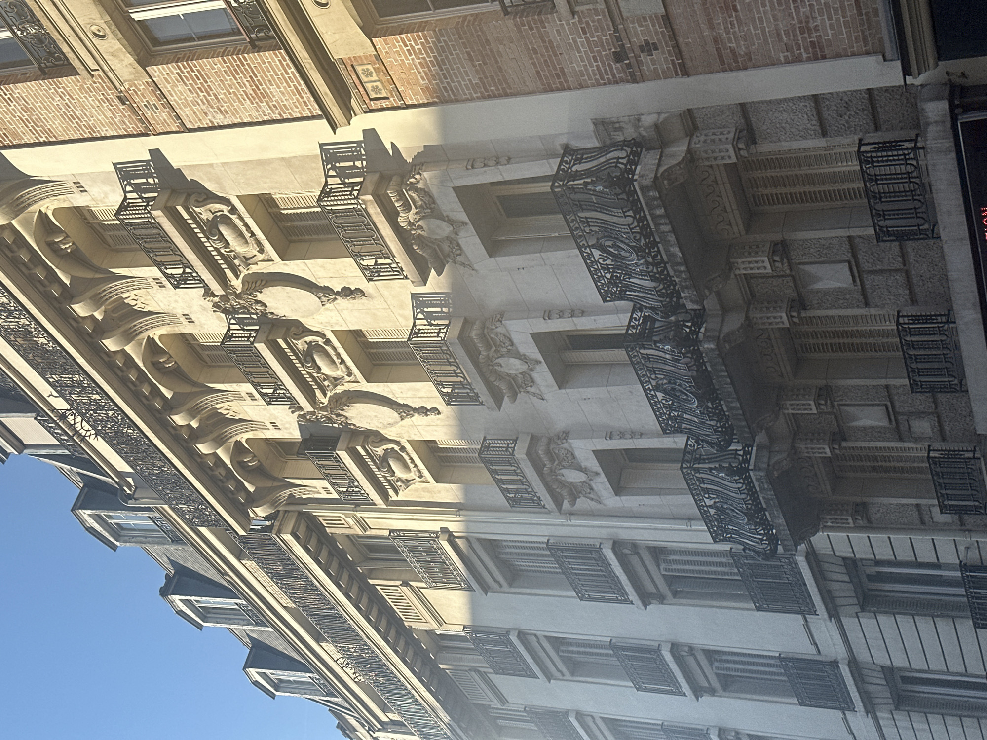 This is a photograph of the facade of a multi-story building in Paris, France, likely from the Haussmannian era, characterized by its ornate architectural details. The building features light-colored stone or stucco on its main facade, with multiple levels of windows and balconies. The balconies are elaborately decorated with wrought-iron railings and decorative stonework. Some windows have shutters. To the right, a portion of an adjacent building with a red brick facade is visible, indicating a dense urban environment. The lighting suggests it is daytime, with sunlight casting shadows and highlighting the architectural features. The sky above is clear and blue. There are no people visible in the frame.
